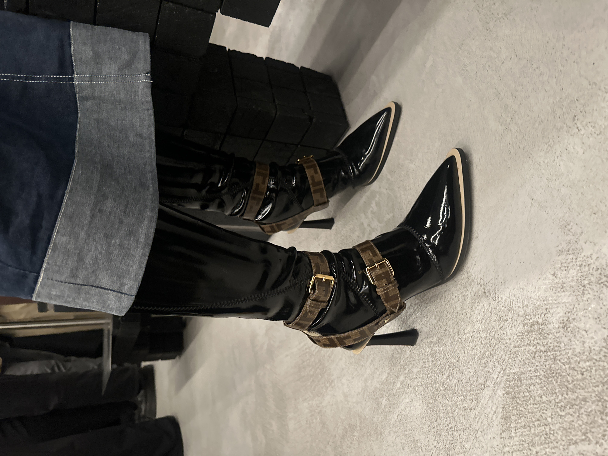 An interior photograph captures the lower legs and feet of an individual wearing black patent leather, pointed-toe, high-heeled boots. Each boot features two brown straps with gold buckles, displaying the Fendi 'FF' monogram, and a light-colored trim around the pointed toe. The subject's dark blue garment, with a wide, light blue denim-style cuff, is visible at the top of the frame. The individual stands on a light grey, textured floor. To the right, a structure composed of stacked black rectangular blocks is present. Blurred lower extremities of other individuals, dressed in dark clothing, are visible in the background to the left. This scene is located at the Fendi boutique, New Bond Street, London, United Kingdom.