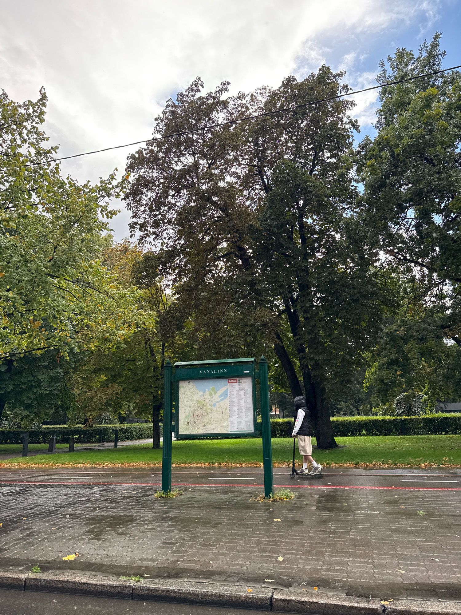 A person is riding a scooter on a wet paved path next to a road with a red line. The person is wearing a black vest over a white long-sleeved shirt, beige shorts, a white helmet, and white sneakers. They are positioned in front of a park with trees and a map display. The map display is green and has the word "VANALINN" at the top, indicating it is a map of Tallinn's Old Town. The weather appears overcast, and the ground is wet, suggesting recent rain. The environment is an urban park setting, likely in Tallinn, Estonia.