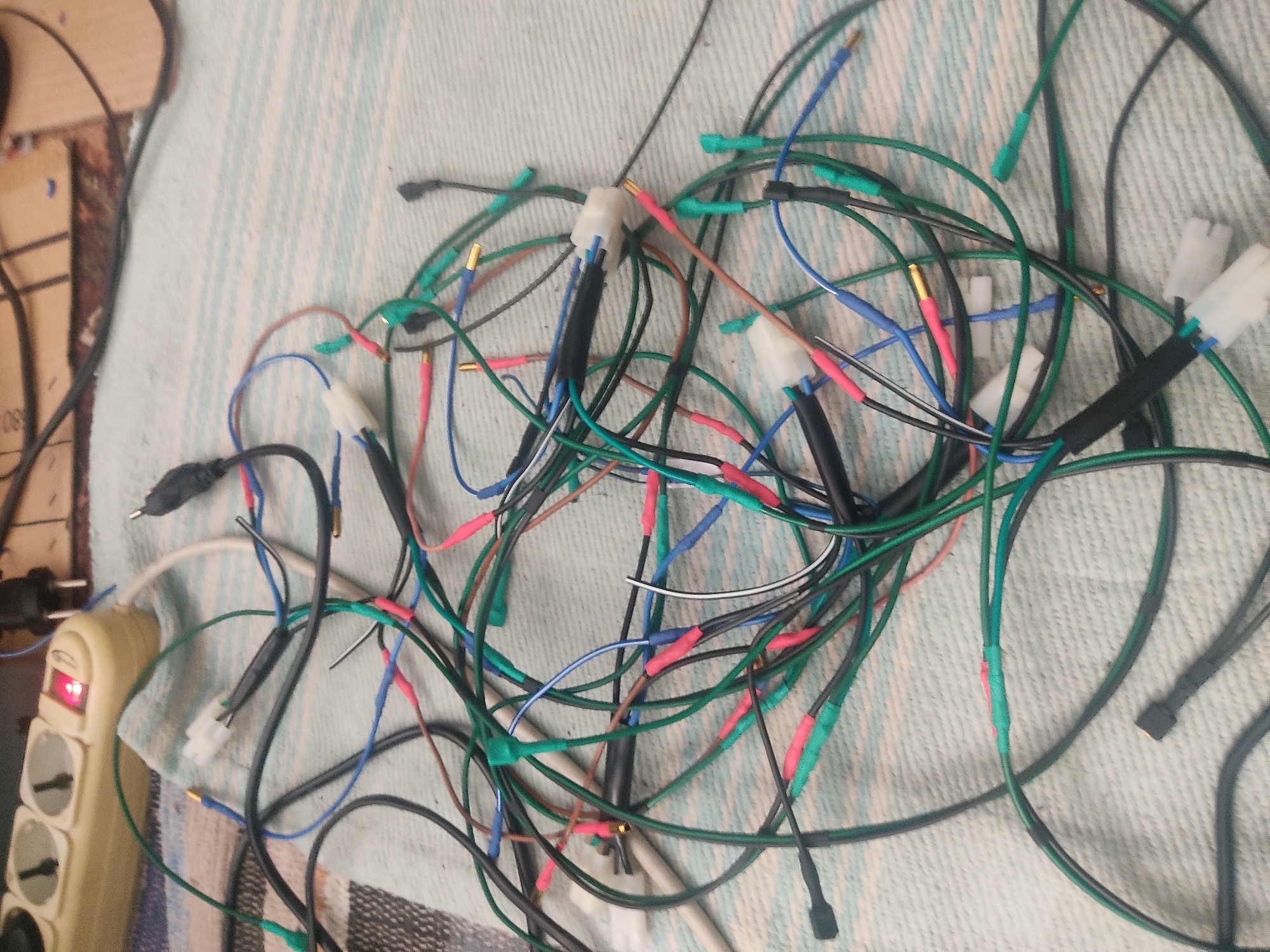 The media file displays a tangled mass of electrical wires, predominantly green, with accents of blue, red, and black. These wires are connected using various methods, including what appear to be heat-shrink tubing, electrical tape, and some with exposed gold connectors. Several white plastic connectors are also visible, suggesting that these wires are part of a larger electronic system or assembly.
In the lower left corner of the image, a beige power strip with multiple outlets is visible. One of the outlets has a red indicator light illuminated. This power strip is connected to a black electrical cord. The wires are spread across a patterned surface, likely a fabric, with light blue and white stripes. The lighting suggests an indoor setting, possibly a workbench or a cluttered desk area. There is a hint of cardboard in the upper left corner, with some faint black markings, possibly numbering like "08". No people are visible in the image. The overall impression is one of a technical or DIY electronics project. The focus is entirely on the wires and the power strip, with no other significant objects or environmental details. The scene is static, with no discernible activity taking place.