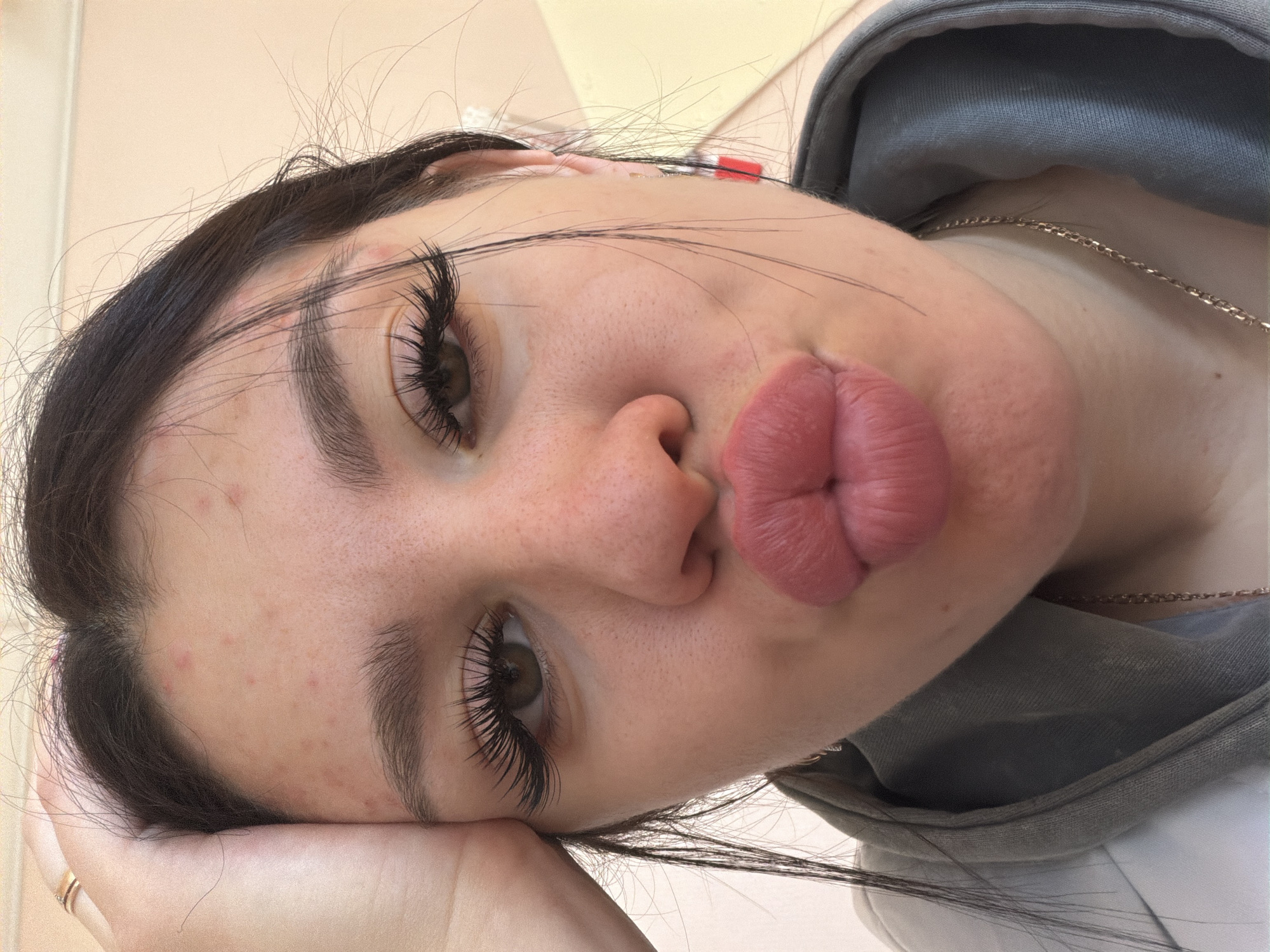 The image is a close-up, forward-facing selfie of a young woman's face. She is leaning her forehead against her left hand, which is resting on her head. Her lips are pursed in a kissing gesture, and her eyes are open. She has long, dark eyelashes, dark eyebrows, and dark hair. Her skin has some small red blemishes, particularly on her forehead. She is wearing a light gray hooded sweatshirt and a thin silver necklace. A red and white earring is visible on her right ear. The background is a plain, light-colored wall. The location is identified as Гаревая, Russia.