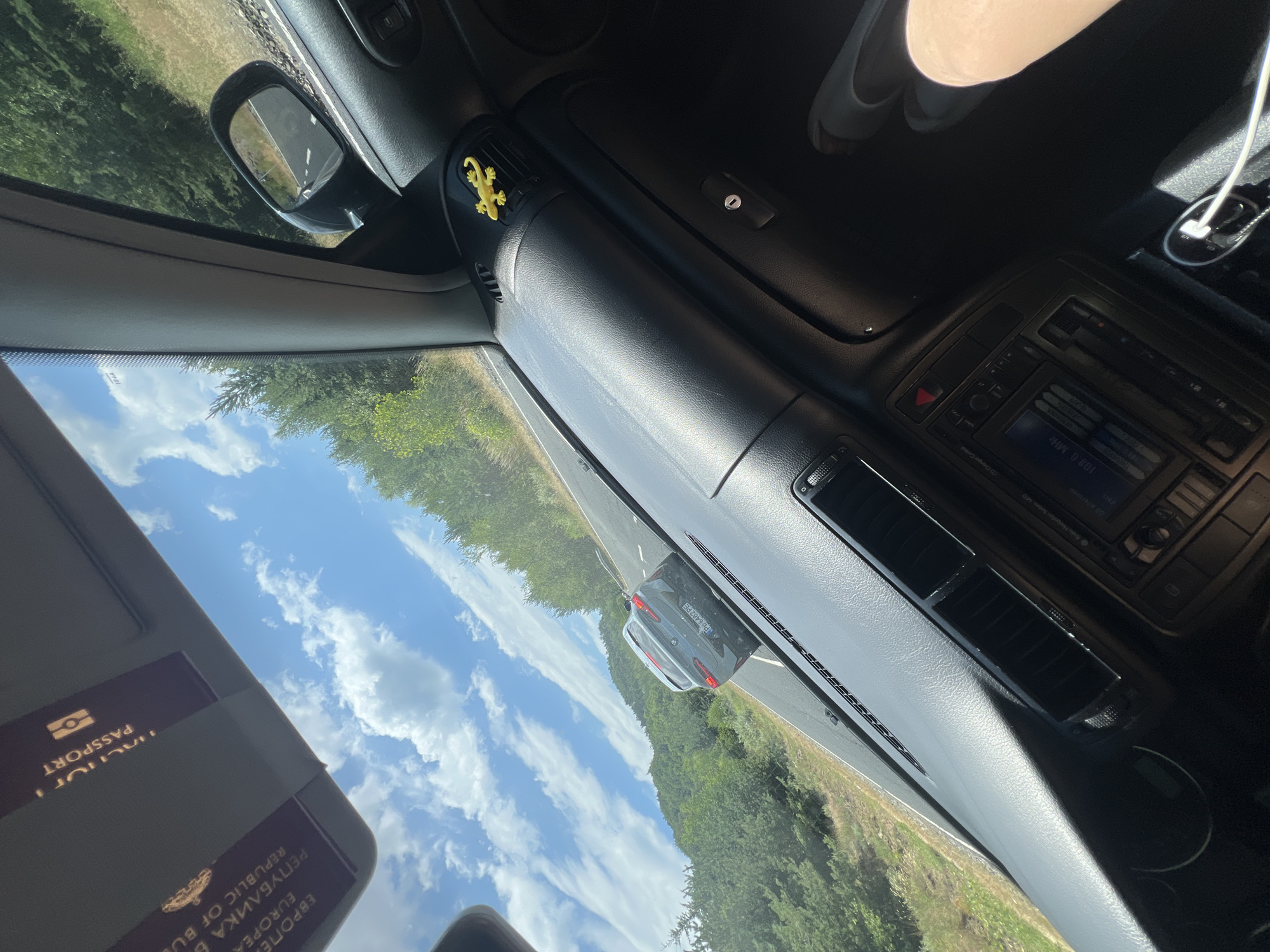 The image is a first-person perspective from inside a car, driving on a road surrounded by lush green trees and hills. The sky is a bright blue with scattered white clouds. In the foreground, the car's dashboard is visible, including the steering wheel, air vents, and a car stereo. A yellow gecko-shaped air freshener is attached to one of the air vents. A passport is visible in the sun visor, showing text in Cyrillic and English, including "PASSPORT" and "REPUBLIC OF BULGARIA". The car is on a paved road, and a gray BMW is in front of it with a Bulgarian license plate. The location is identified as Lozengradtsi, Bulgaria. The scene is captured during daylight.
