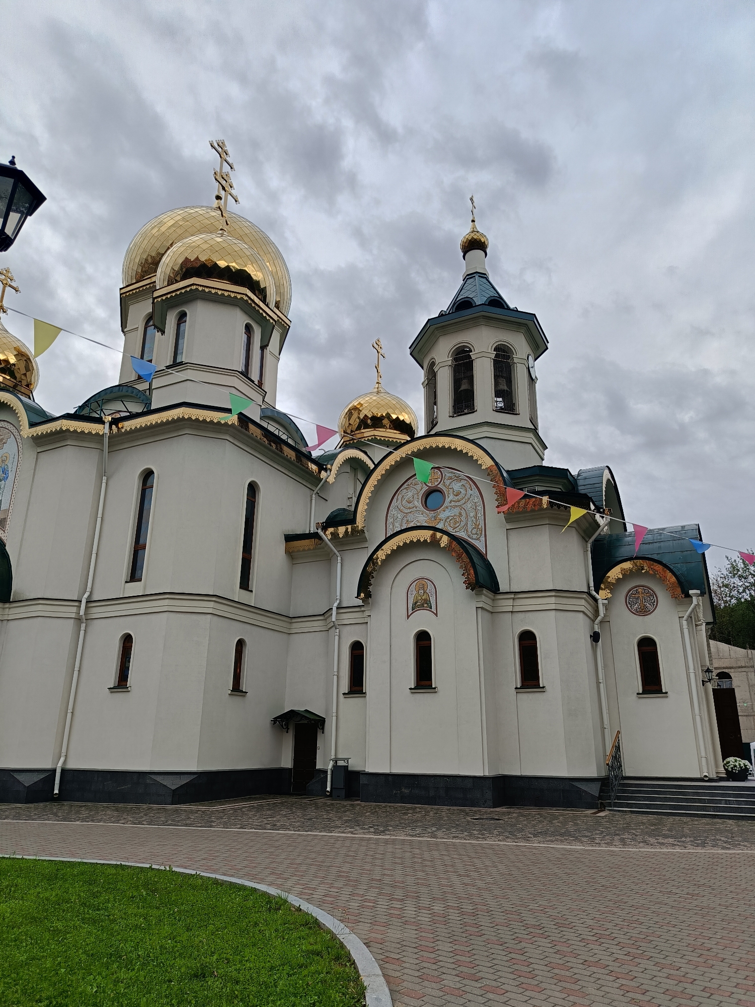 This image depicts an Orthodox church, likely in Saint Petersburg, Russia, under a cloudy sky. The church is a prominent white building with multiple golden domes topped with crosses. Several smaller domes and decorative elements also feature gold accents. Small, colorful triangular flags are strung across the upper parts of the building, suggesting a festive occasion or celebration. The architecture includes arched windows, ornate detailing above some of the window arches, and a bell tower with a blue roof. The foreground shows a paved walkway made of brick-like stones, with a curved patch of green grass to the left. The overall impression is of a grand religious structure on a somewhat overcast day. No people are visible in the image, and there is no discernible text.