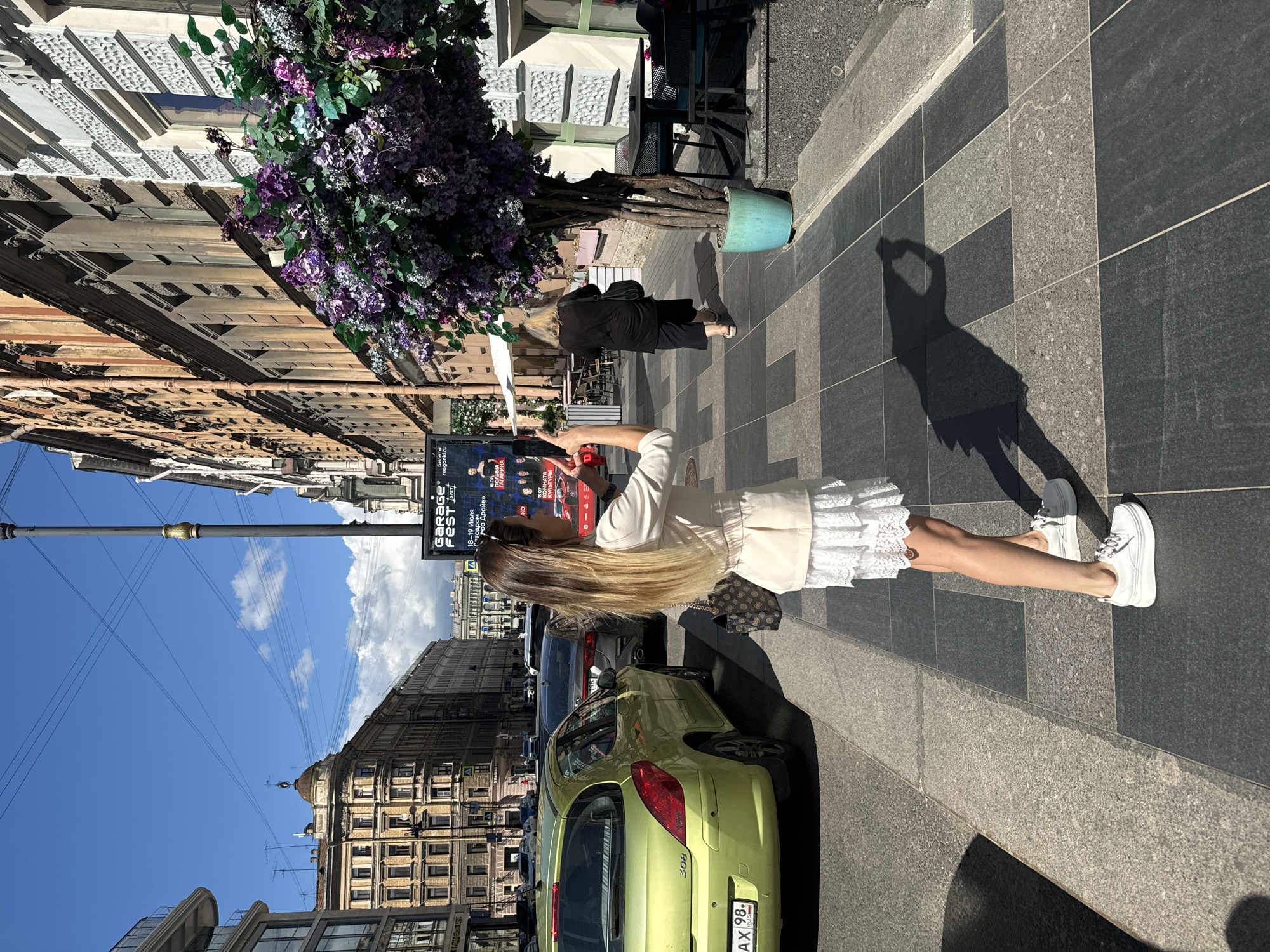 A woman in a light-colored jacket and white lace skirt is standing on a sidewalk in Saint Petersburg, Russia, holding a phone up to take a picture. She is wearing white sneakers and carrying a brown patterned bag. A lime green car is parked on the street next to her. In the background, there are buildings with ornate architecture and a large tree with purple flowers in a turquoise pot. A poster for "GARAGE FEST" is visible on the right side of the image. The sky is blue with some clouds.