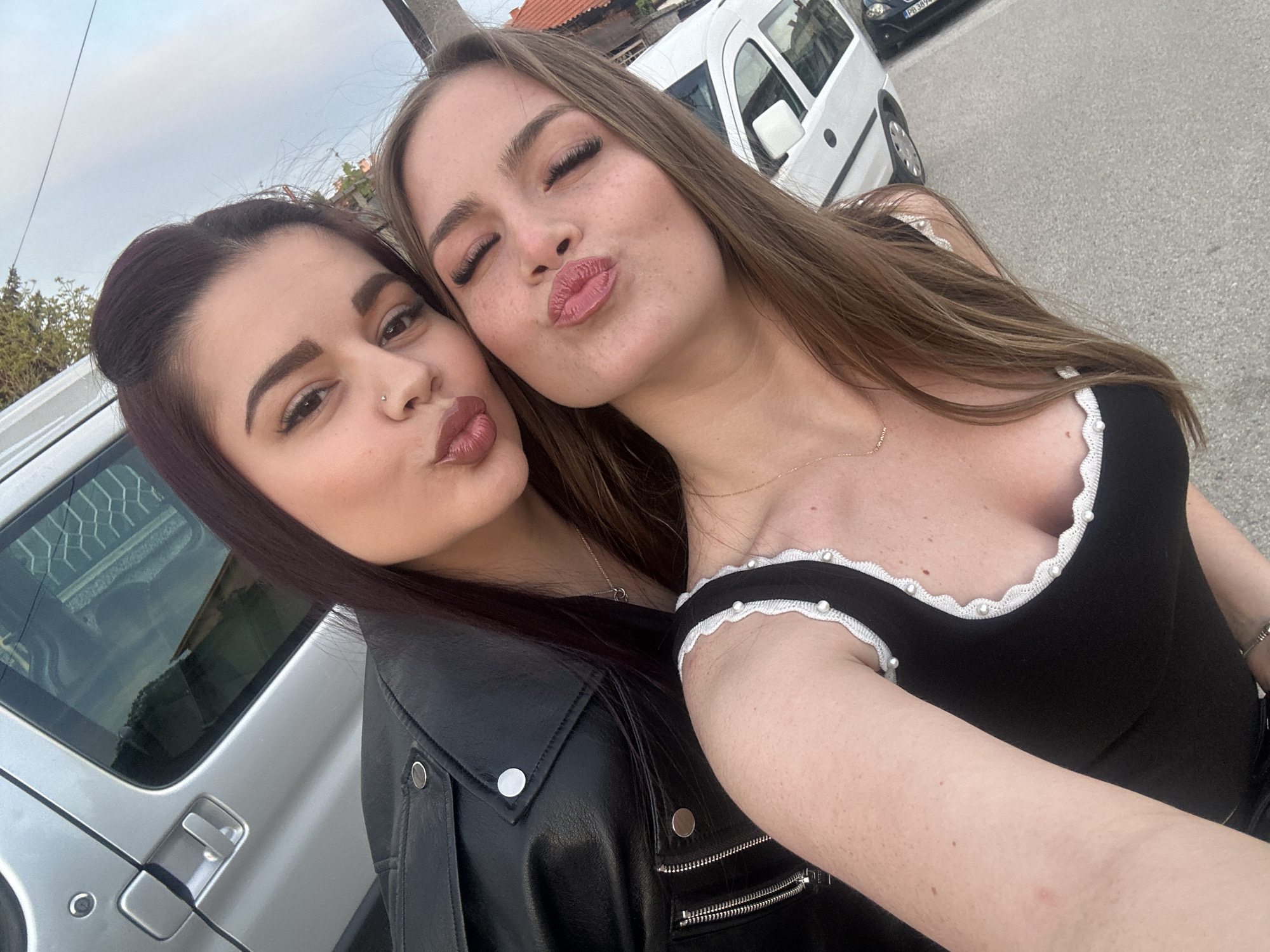 Two young women are posing closely for a selfie, both puckering their lips in a kissing gesture. The woman on the left has dark hair pulled back, wears a black leather jacket, and has a nose piercing. The woman on the right has long, light brown hair and a black top with white trim. Behind them is a street scene. A white van and a silver SUV are parked. The image was taken in the city of Parvomay, Bulgaria.