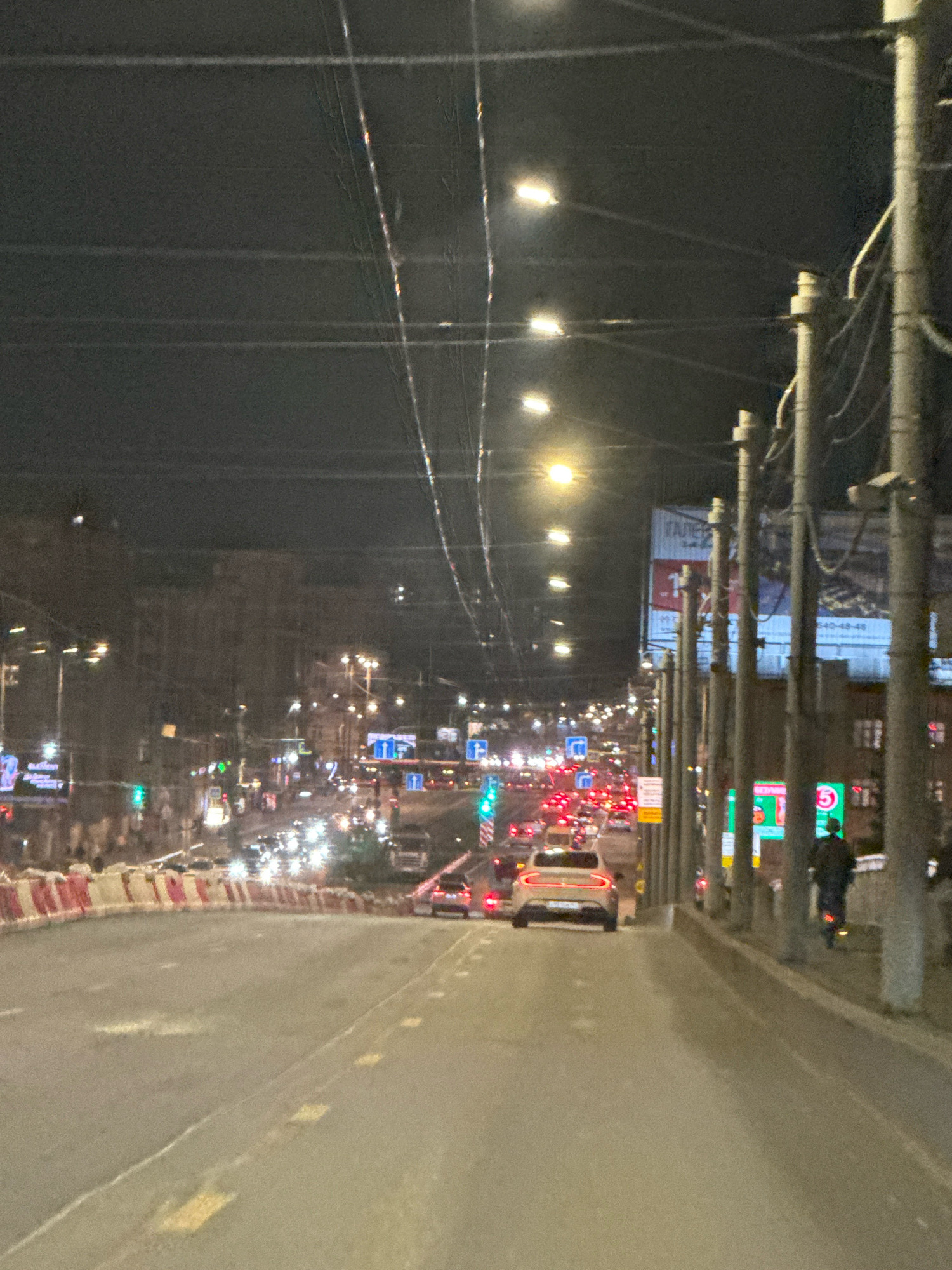 A nighttime street scene in Saint Petersburg, Russia, shows a busy road with traffic. Multiple lanes of vehicles are visible, with headlights and taillights creating streaks of light. Streetlights illuminate the road and the surrounding buildings, which appear to be urban architecture. Overhead wires and utility poles are prominent features of the scene. A person is riding a scooter on the sidewalk. Road barriers are present along the side of the road. Billboards and signs are visible on buildings. The overall impression is of a bustling city at night.