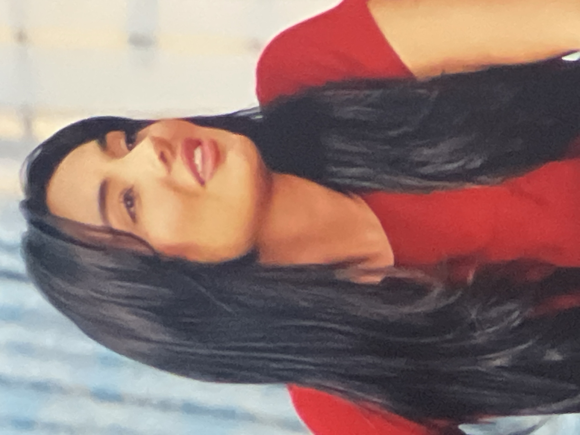 The image is a medium shot of a woman with long, dark hair, wearing a red top. She is looking to her right and her mouth is slightly open. The background is out of focus and appears to be a room with light-colored walls and some blue elements. There is no indication of the city of Vanadzor, Armenia, or any specific urban environment.