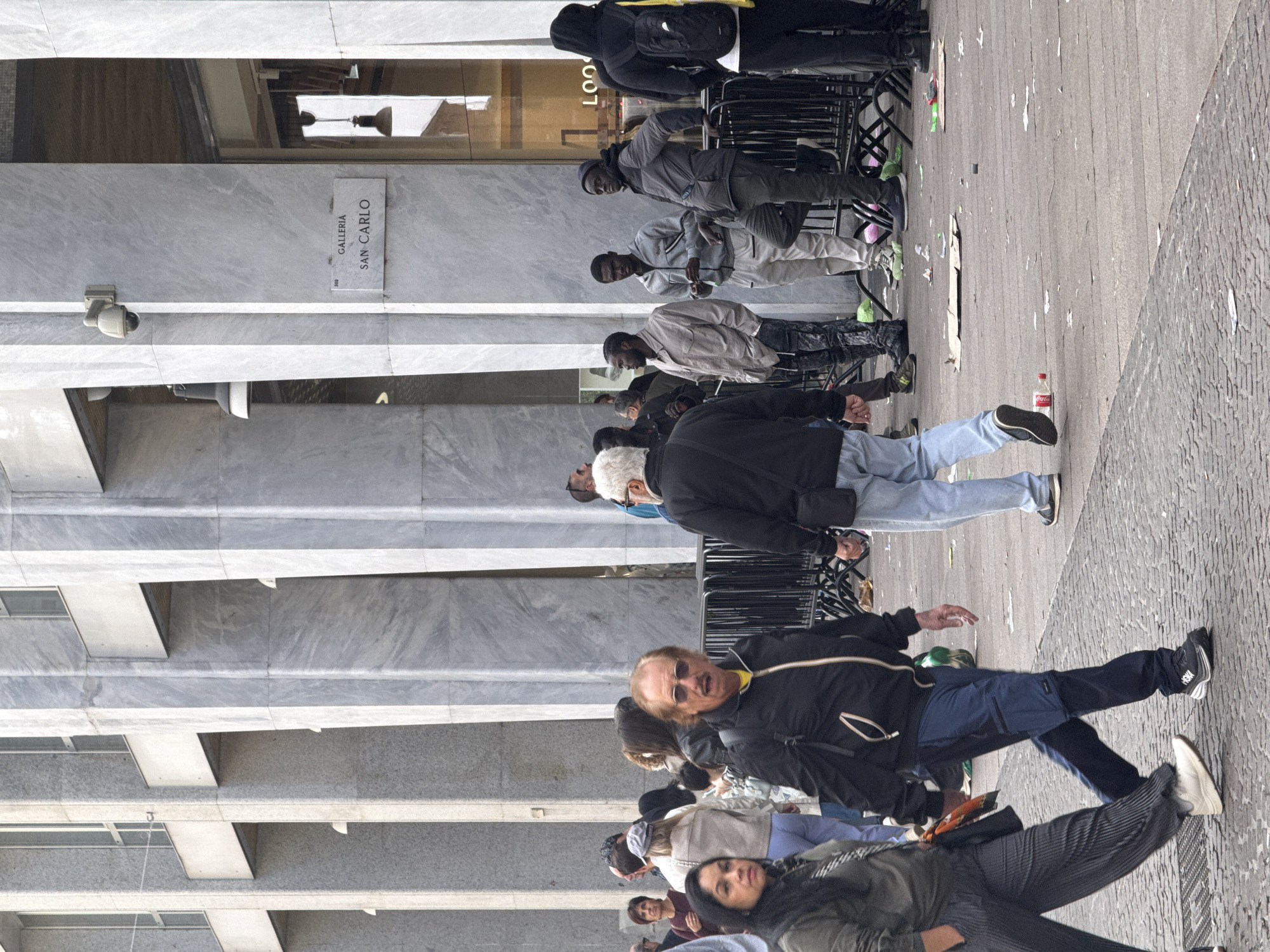 The image depicts a street scene in Milan, Italy, with people on the sidewalk in front of a building. A sign on the building reads "Galleria San Carlo." Several people are visible, including a group of men standing near a temporary barrier with green sneakers, and a couple walking towards the camera in the foreground. The ground is littered with trash. The architectural style of the building is modern.
