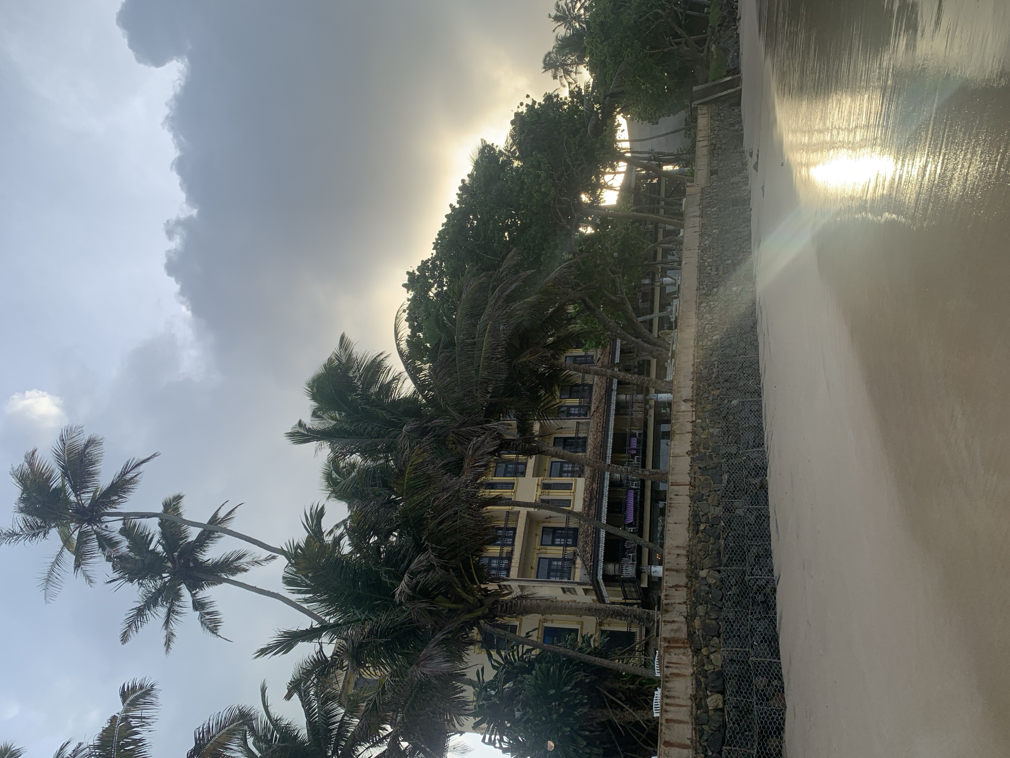 This is a photograph of a beachfront in Kathaluwa, Sri Lanka, at what appears to be either sunrise or sunset, given the low angle of the sun and the reflection on the wet sand. In the background, a multi-story building with a yellow facade and dark windows is visible, framed by numerous tall palm trees. The building has balconies with dark railings and a tiled roof section. A stone retaining wall, reinforced with wire mesh cages filled with rocks, separates the beach from the building's grounds. The foreground is dominated by a sandy beach with the tide line marked by wet sand reflecting the sky. The sky is overcast with dramatic clouds, but a patch of brighter sky suggests the sun is near the horizon, creating a lens flare effect on the water's surface. There are no people or animals visible in the image.