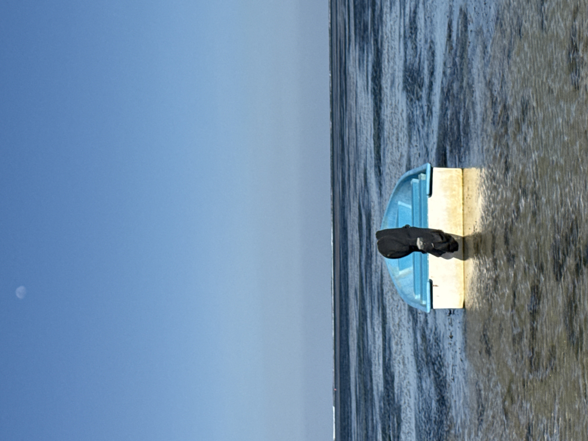 The image depicts a small, blue and white boat partially submerged in shallow water. A black garment, possibly a jacket or wetsuit, is draped over the stern of the boat. The water is calm and reflective, with ripples indicating a gentle current. In the distance, a thin horizon line separates the water from a clear blue sky. A faint crescent moon is visible in the upper left quadrant of the sky. The scene appears to be a tranquil coastal environment. The image does not contain any elements that suggest a specific location within Denpasar, Indonesia.