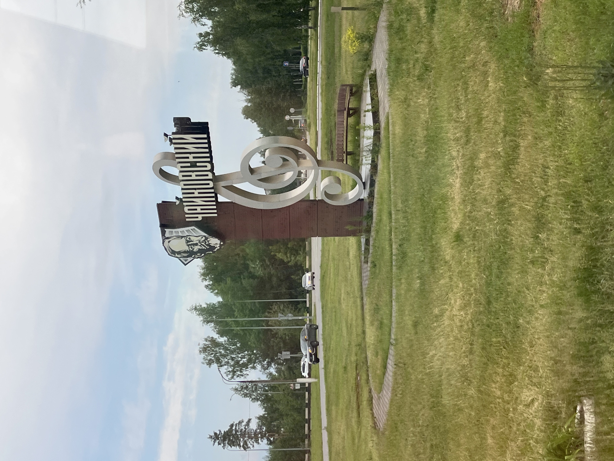 The image captures an outdoor scene, likely a public park or roadside area, in Chaikovsky, Russia. The central feature is a prominent monument or welcome sign dedicated to the city and its namesake, Pyotr Ilyich Tchaikovsky.
The sign consists of several elements: a vertical black block displaying the text "ЧАЙКОВСКИЙ" (Chaykovskiy or Tchaikovsky) in white Cyrillic letters. Below the text, a black-and-white stylized portrait, presumably of Pyotr Ilyich Tchaikovsky, is visible. To the right of the text, a large, ornate musical treble clef (G-clef), crafted from a light-colored material, stands tall. The entire structure is supported by a dark brown base.
To the right of the monument, a manicured grassy lawn with a curved, light-colored pedestrian pathway extends into the distance. A wooden park bench is situated near the sign, beside the pathway. In the midground, a paved road with several cars, including white and dark-colored vehicles, is visible, indicating ongoing traffic. Streetlights and utility poles line the road. The background is filled with dense green trees, suggesting a forested or park-like setting. The sky is partly cloudy, with light blue and white hues, indicating daytime. The scene conveys a sense of civic pride and commemoration.