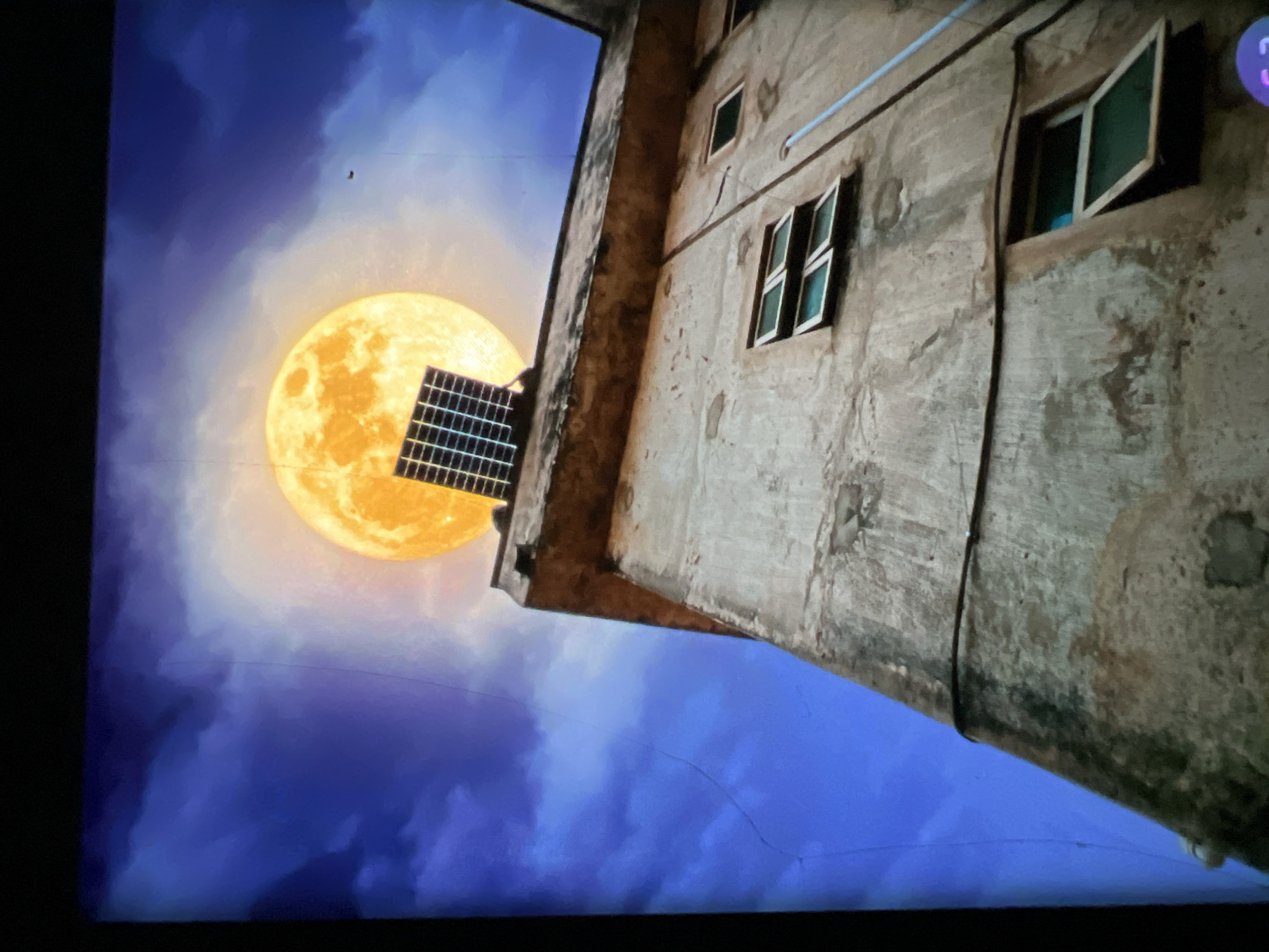 A low-angle shot captures a weathered, concrete building against a vibrant, cloudy sky with a large, luminous full moon. A solar panel is positioned on the edge of the building's roof, silhouetted against the moon's glow. The building features multiple windows, some open and some closed, on its textured facade. The scene appears to be an artistic or surreal depiction, possibly a composite image, rather than a literal photograph of a specific location in Lagos, Nigeria. The image does not contain any people or discernible actions or interactions.