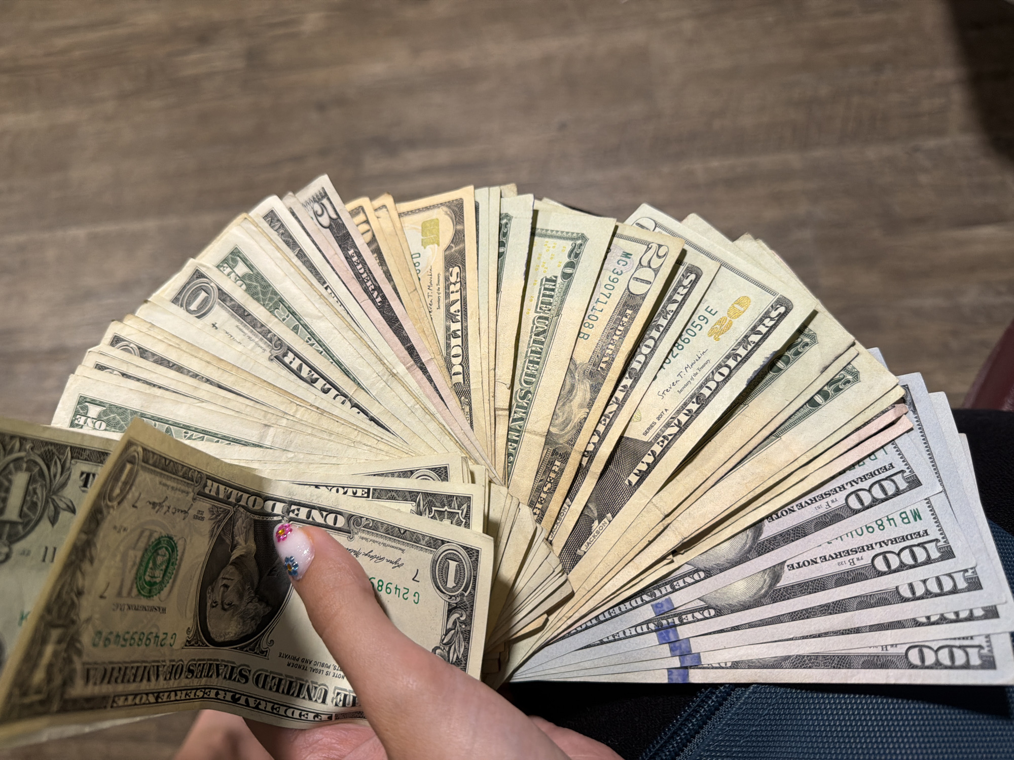 A hand with a decorated fingernail is holding a fanned-out collection of U.S. dollar bills against a wooden surface. The bills appear to be of various denominations, including $1, $5, $10, $20, and $50. Some of the bills show clear serial numbers and the text "FEDERAL RESERVE NOTE" and "THE UNITED STATES OF AMERICA." The image is taken from a top-down perspective. There is no indication of the city of Lake Delton, Wisconsin, or any specific location within it.