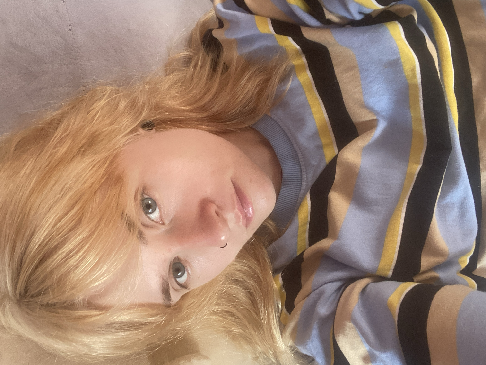 A young woman with blonde hair and blue eyes is lying down, looking at the camera. She is wearing a striped shirt with blue, tan, black, and yellow stripes. She has a nose ring. The background appears to be a light-colored surface, possibly a bed or couch. The image is taken from a slightly overhead angle. The location is not identifiable as Chaikovsky, Russia.