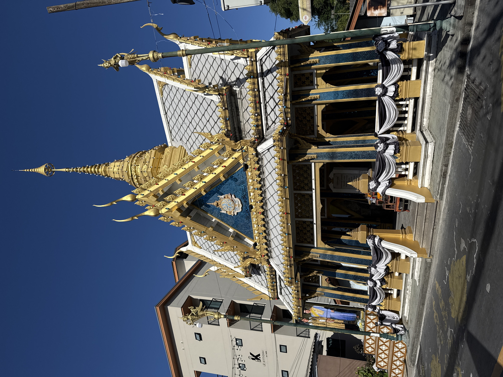 In Patong, Thailand, a small, ornate Buddhist temple with a golden spire and intricate blue and gold facade stands prominently. The temple's roof is adorned with decorative golden finials, and a crest with blue and gold detailing is centered above the entrance. Two life-sized statues of people in traditional attire are placed to the left of the steps leading to the temple. The temple is decorated with draped black and white fabric, suggesting a period of mourning or a special ceremony. To the left of the temple, a modern, multi-story building with a sign that reads "LE KITTICHAI" is visible. A street lamp with a golden mythical creature figurine is positioned in front of the temple. The scene is set against a clear blue sky. The street in front of the temple is paved with asphalt and marked with yellow and white lines.