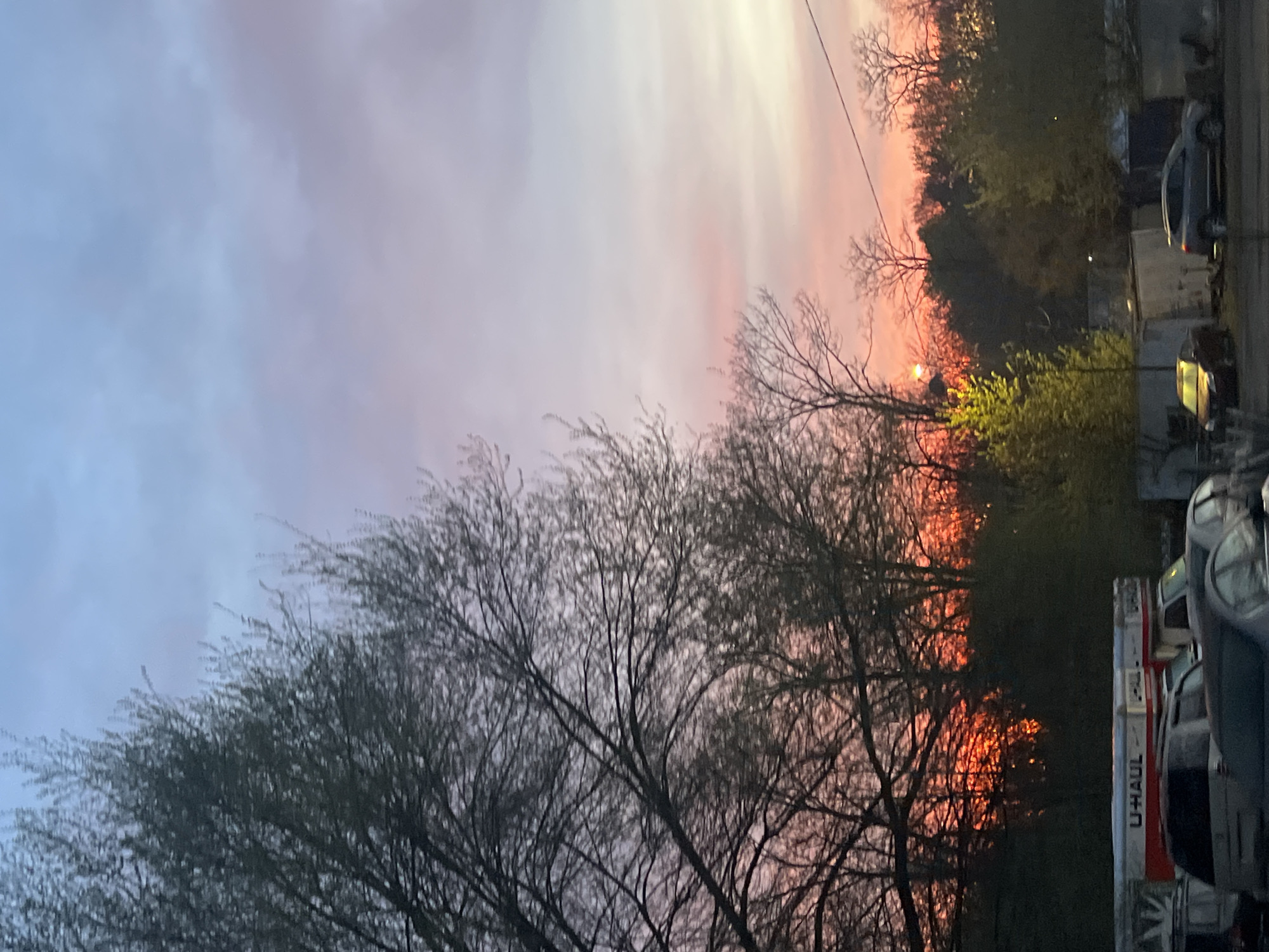The image presents a landscape view, seemingly in the early evening or dawn. A large tree with bare branches dominates the foreground. Beyond the tree, there's a mix of other trees and bushes, creating a layered effect. The sky displays a gradient of colors, ranging from a pale lavender-gray at the top to a more vibrant pink and orange near the horizon, indicative of a sunrise or sunset. In the background, on the right side of the frame, parked vehicles are visible. Based on the presence of a U-HAUL truck, the location can be inferred to be in proximity to a rental or moving service in the City of Newburgh, United States of America. The setting appears to be urban or semi-urban, given the presence of vehicles and what looks like structures behind the trees. There are no people visible in the image.