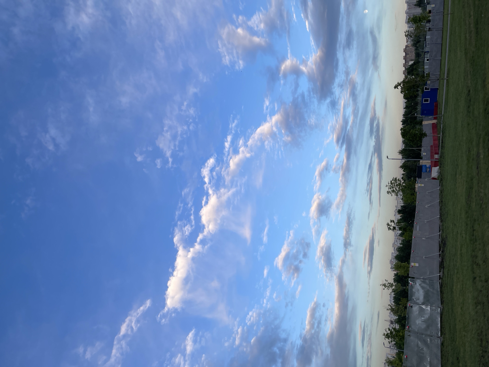 This is a vertical, wide-angle photograph of a sky with clouds over a grassy area with a fence and some buildings in the distance. The sky is a vibrant blue with scattered white and gray clouds. The clouds range from wispy cirrus to larger, more dense formations, some tinged with pink and orange from the setting sun.
The ground is covered in green grass. A metal fence, made of modular panels, runs horizontally across the lower right portion of the image. Behind the fence, there are trees and then a line of buildings, suggesting an urban or suburban setting. A small blue structure with a white square window is visible between the trees and the fence. A streetlamp is also visible near the fence.
The time of day appears to be late afternoon or early evening, given the quality of the light and the colors of the clouds. The weather seems to be clear, with some cloud cover. The location is Moscow, Russia. No people are visible in the frame. There is no discernible text in the image.