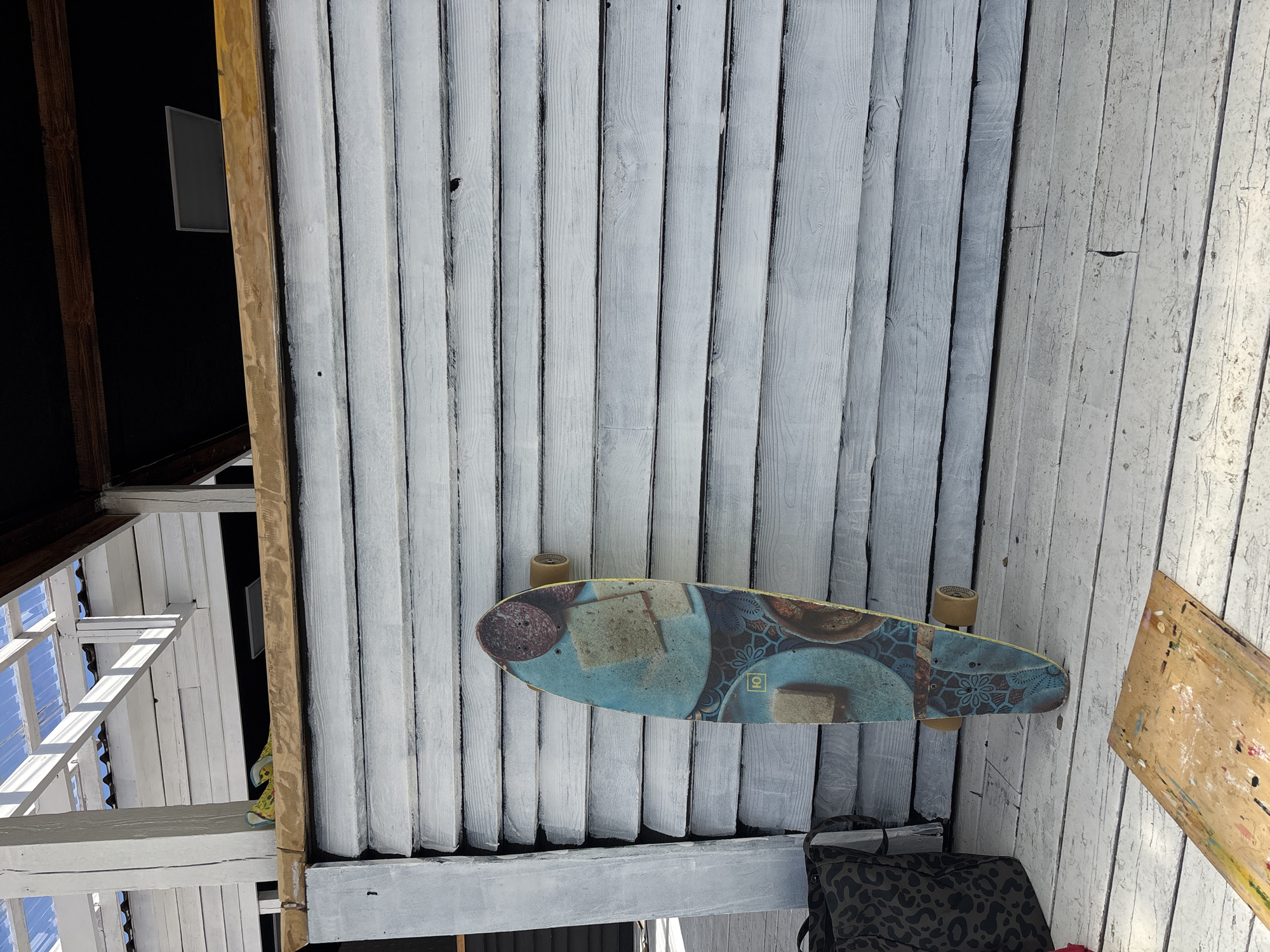 The image is presented rotated 90 degrees counter-clockwise, depicting what appears to be an interior or covered exterior ceiling constructed from wooden planks, painted light gray or white, with visible wear and dark lines between them. A longboard or skateboard with two wheels visible is positioned horizontally against these planks. Its underside features a colorful graphic with a blue background, intricate patterns, and depictions of round objects (resembling sliced salami) and square objects (like cheese). A small square logo with an 'O' shape is visible on the skateboard.
To the left, a dark area, possibly an adjoining room or void, is framed by a light brown wooden beam. Within this dark space, a light gray rectangular object, perhaps a monitor or mirror, is mounted. In the lower left quadrant, natural light streams through an opening, illuminating white wooden structural beams and revealing a clear blue sky through what appears to be a skylight or high window.
On the right side, a wooden artist's palette, stained with various colors of paint, leans against the wooden planks. Below the skateboard, a black bag or backpack with a leopard print pattern is partially visible. The scene is brightly lit, indicating daytime and sunny weather. No people are present, and no activity is taking place. The setting is in Дрокино, Russia.