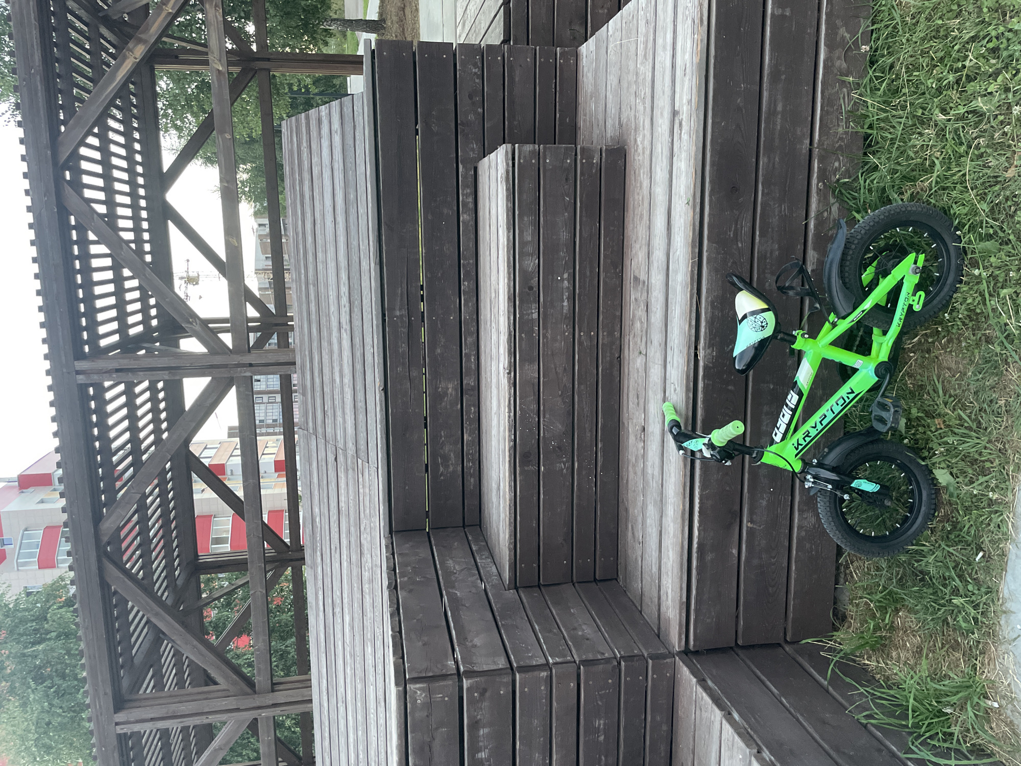 A bright green Krypton bicycle with neon green handlebars is parked on the grass in front of a wooden tiered structure. The bicycle has black tires and a multi-colored seat. The wooden structure appears to be an outdoor seating area or amphitheater with dark brown wooden planks. Behind the wooden structure, a modern building with red and white accents is visible, along with trees and a construction crane in the distance. The scene suggests an outdoor recreational area in an urban or suburban setting. This image was taken in the city of Laishevo, Russia.