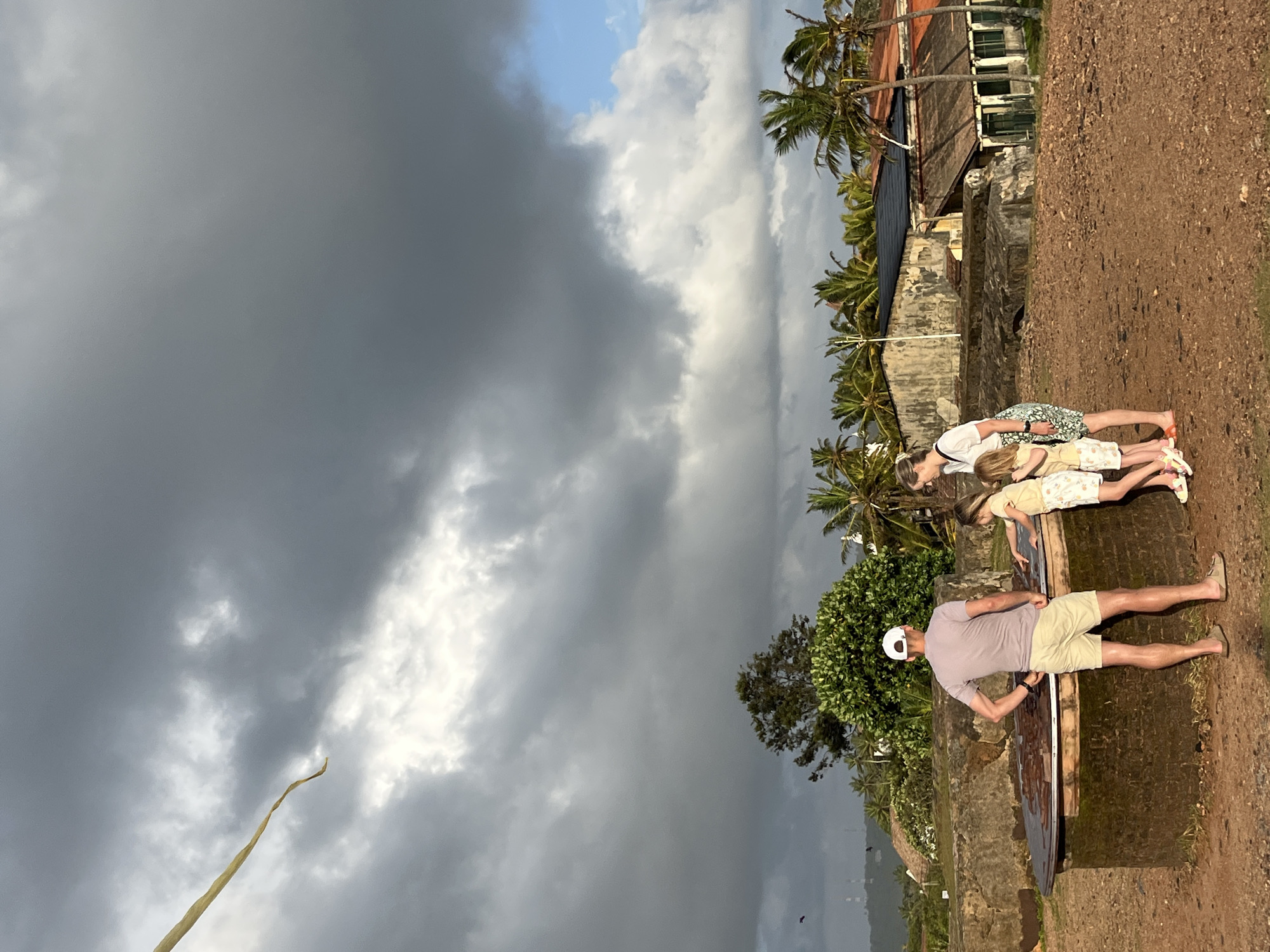 In Galle, Sri Lanka, a family stands near a circular, low-lying structure with a textured surface, possibly a historical artifact or exhibit, in the foreground. The man, wearing a white cap, a light purple t-shirt, and beige shorts, stands with his back to the camera, facing the structure. A woman in a white t-shirt and patterned skirt stands to his right, looking down at the structure. Two young girls, one in a yellow top and patterned shorts and the other in a white top and patterned shorts, are also gathered around the structure, interacting with it. They are all on a dirt path. Behind them, a wall made of large stones is visible. Beyond the wall, lush green trees and palm trees are abundant, and in the distance, several buildings with terracotta roofs and weathered facades are visible. The sky is dramatic, with a mix of dark, heavy storm clouds and patches of bright blue sky, suggesting changing weather. A long, pale yellow streamer or kite tail hangs from the upper left of the frame against the stormy sky.