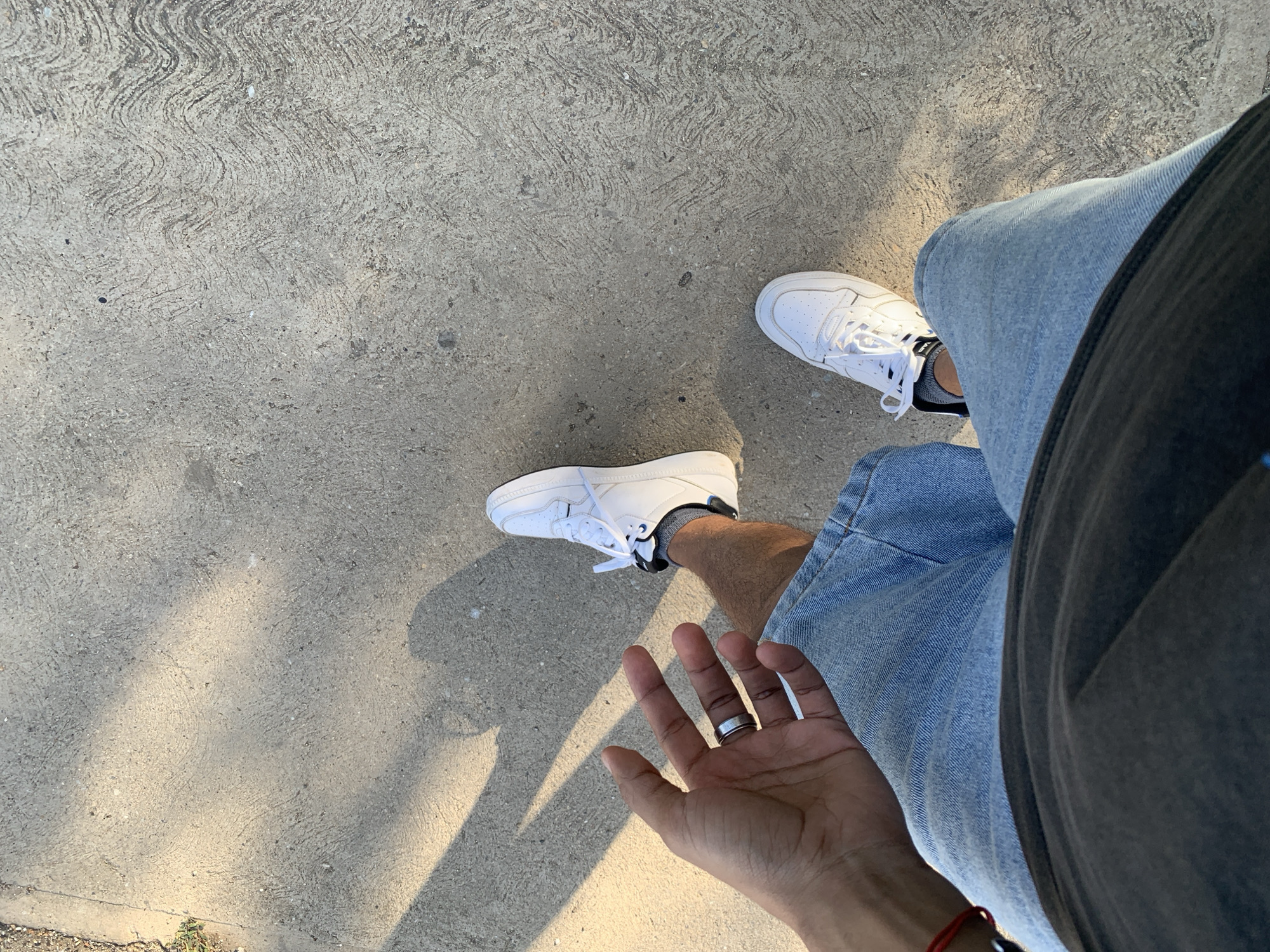 The image is a first-person perspective shot of a person standing on a concrete surface outdoors. The person is wearing white sneakers, denim shorts, and a dark t-shirt. Their left hand, adorned with a ring, is extended towards the camera. Sunlight casts shadows across the concrete, which has a textured, wavy pattern. There is no visible indication that this scene is located in El Valle del Espíritu Santo, Venezuela.