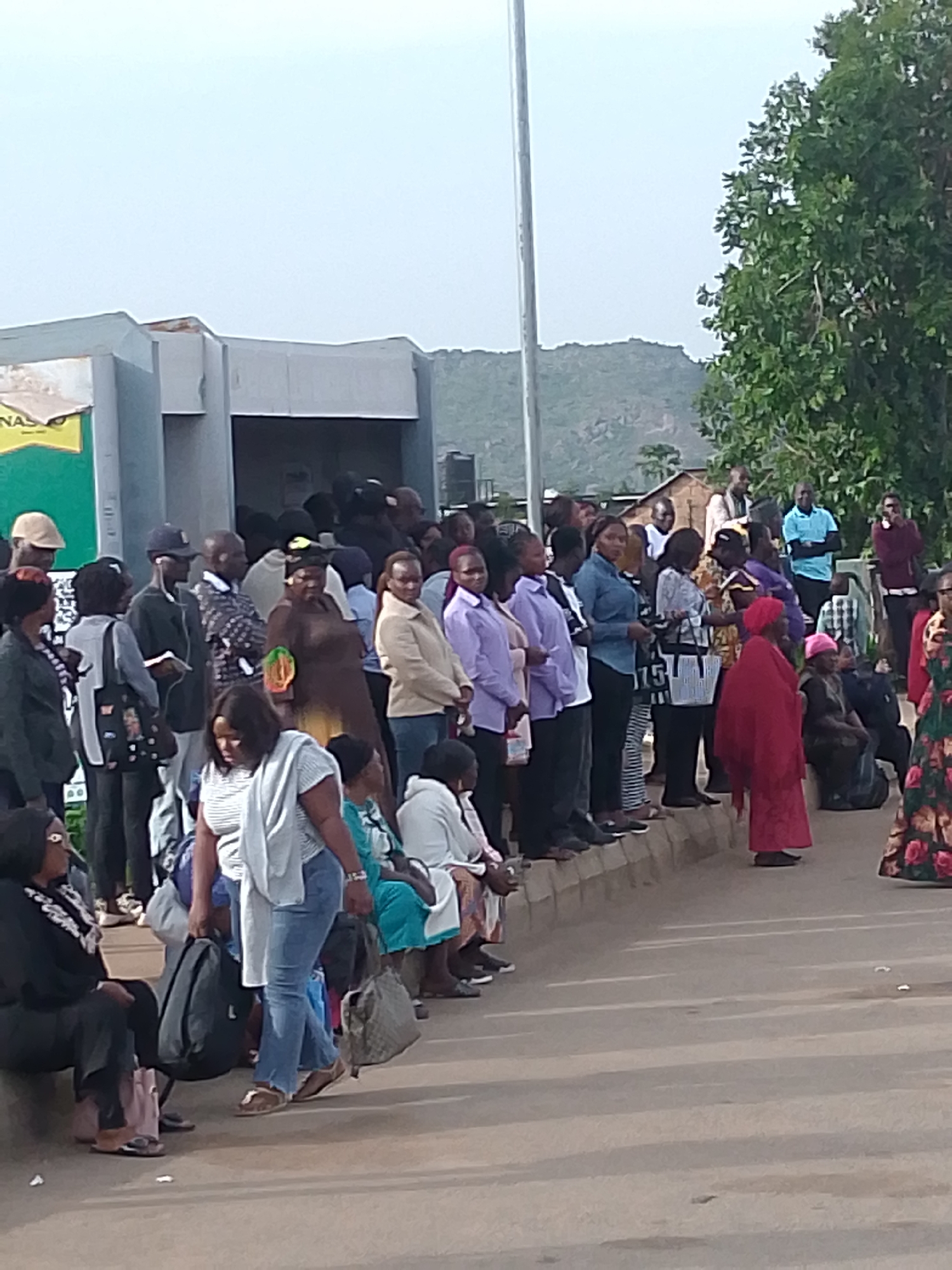 A group of people are gathered outside a building, with some standing and others sitting. The building appears to be a low-rise structure with a light-colored facade. There are trees and a hilly landscape in the background. The individuals are dressed in casual clothing, and some are carrying bags. The scene suggests a public gathering or waiting area, possibly at a transportation hub or a community center. The ground is paved, and there are some shadows indicating the presence of sunlight. The overall atmosphere seems calm and orderly. Based on external knowledge, this image is located in Kuru Dungus, Nigeria.