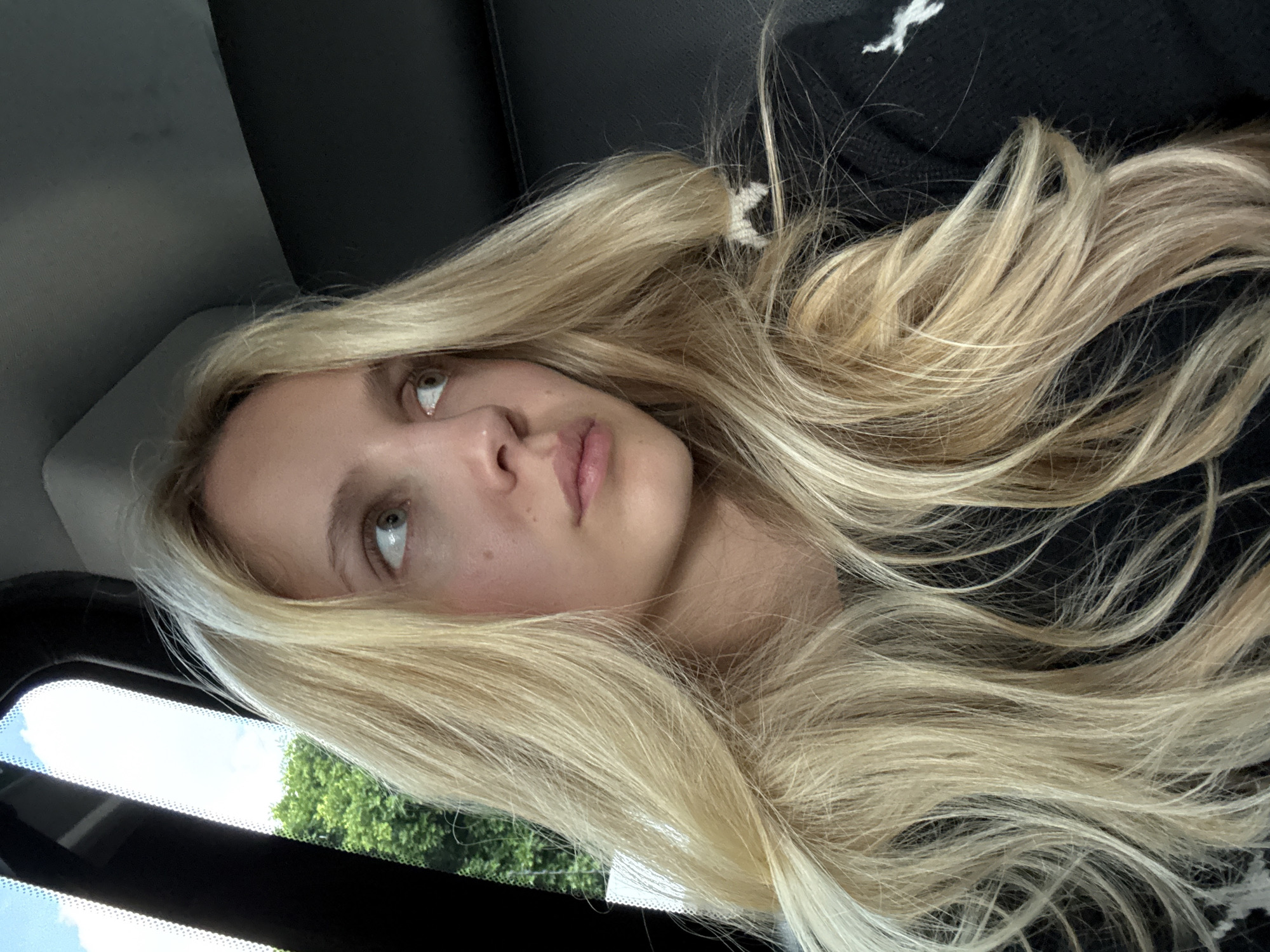 A young blonde woman is captured in a close-up, shoulder-level portrait. She is looking to her right, away from the camera, with a neutral expression. Her long, blonde hair is styled loosely, framing her face. She is wearing a dark sweater with a white design. The background is dark and appears to be the interior of a vehicle. Through a window on the left side of the frame, lush green trees and a bright blue sky with white clouds are visible. The lighting suggests it is daytime. The image does not provide any information to determine the location in Mexico City, Mexico.