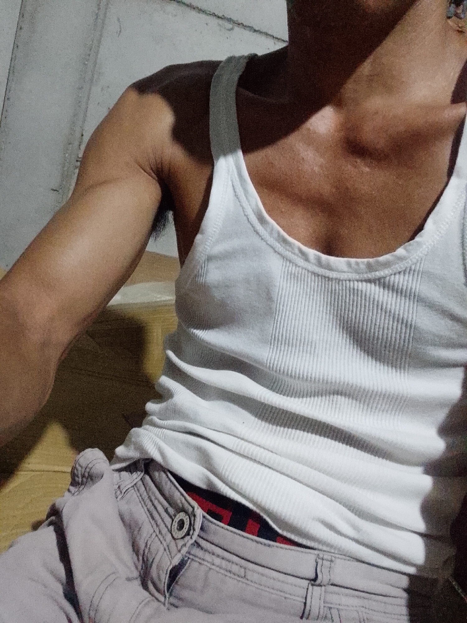 A close-up shot depicts a man's torso and part of his arm, likely taken indoors, possibly in a private residence in Bukit Ketok, Indonesia, though there is no definitive environmental indicator to confirm this. He is wearing a white ribbed tank top. His left arm is visible, showing some muscle definition. He is wearing gray pants with a button closure, with a glimpse of red and black patterned underwear showing above the waistband. The lighting is somewhat dim and uneven. The background appears to be a plain wall.
