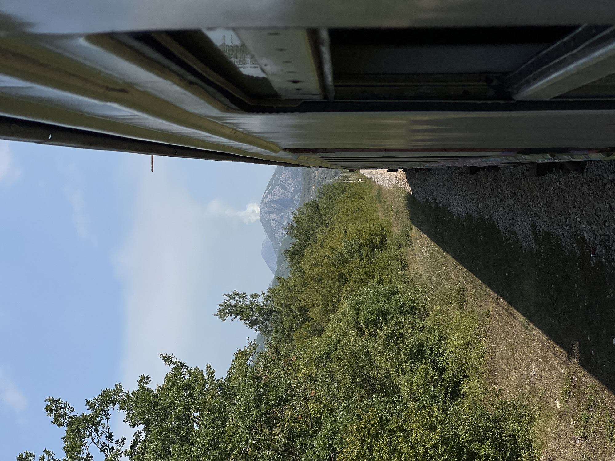 A side view from a moving train shows a plume of smoke rising from a mountain peak in the distance. The train car on the right is light green with visible windows and corrugated siding. To the left of the tracks, dense green foliage lines the side of a hill, with a few trees scattered throughout. The sky is a clear blue with a few wispy clouds. The location is the city of Limljani, Montenegro.