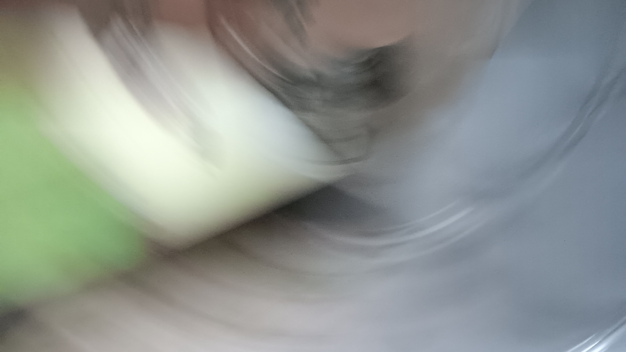 The image is an abstract, motion-blurred depiction of an indoor scene. A large, amorphous, light-colored object, possibly a piece of furniture or fabric, dominates the center-right of the frame, with a distinct greenish hue on its right edge. Swirling grey and white lines suggest movement, perhaps of light or a reflective surface, creating a dynamic, distorted effect across the entire image. The overall impression is one of blurred motion and indistinct shapes, making it impossible to identify specific objects or context. There are no discernible details about people, actions, or the environment that would allow for localization in Sonobekel, Indonesia, or any other specific location.