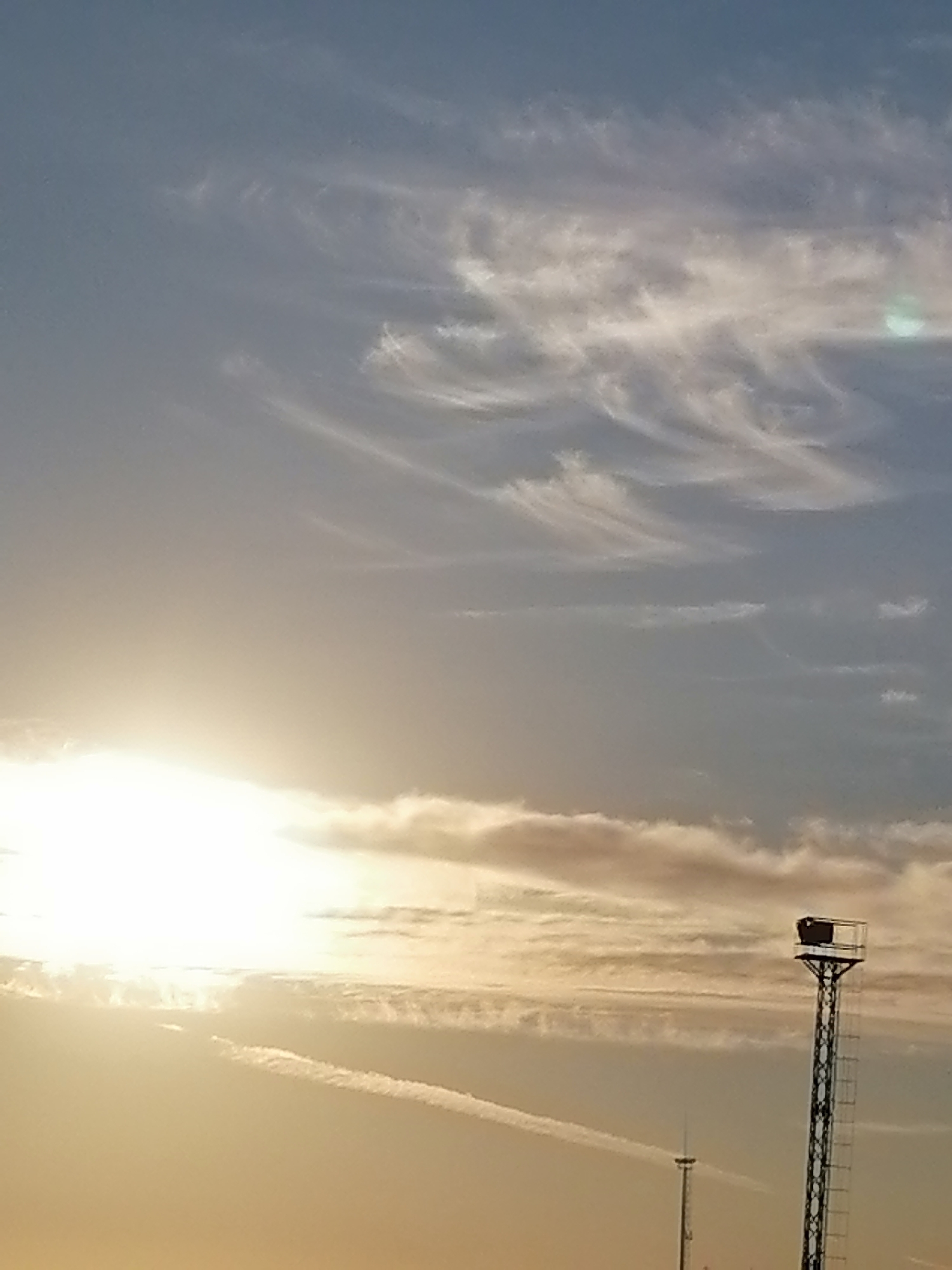 The image depicts a sky with scattered clouds, illuminated by the sun's bright glow on the left side. In the foreground, a tall, lattice-like metal tower with a rectangular structure at its top stands to the right. Further in the background, another smaller tower is visible. The sky transitions from a bright, warm hue near the horizon to a cooler blue at the top. The overall scene suggests an outdoor environment, possibly at dawn or dusk, given the lighting. The location is identified as Kharino, Russia.
