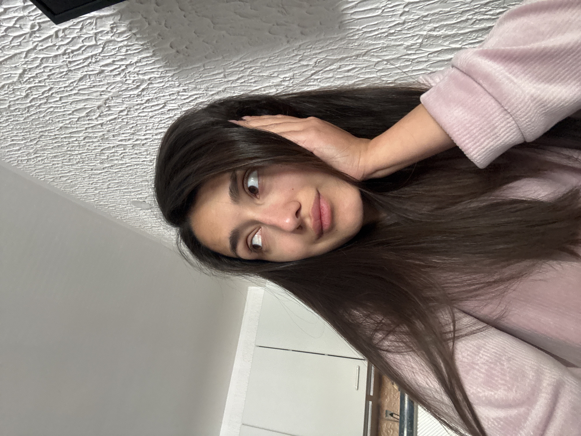 A young woman with long, dark hair is holding her hand to her head. She is wearing a pink sweatshirt. The background includes a textured ceiling and a gray wall with white cabinets. The location is not specified or discernible from the image.