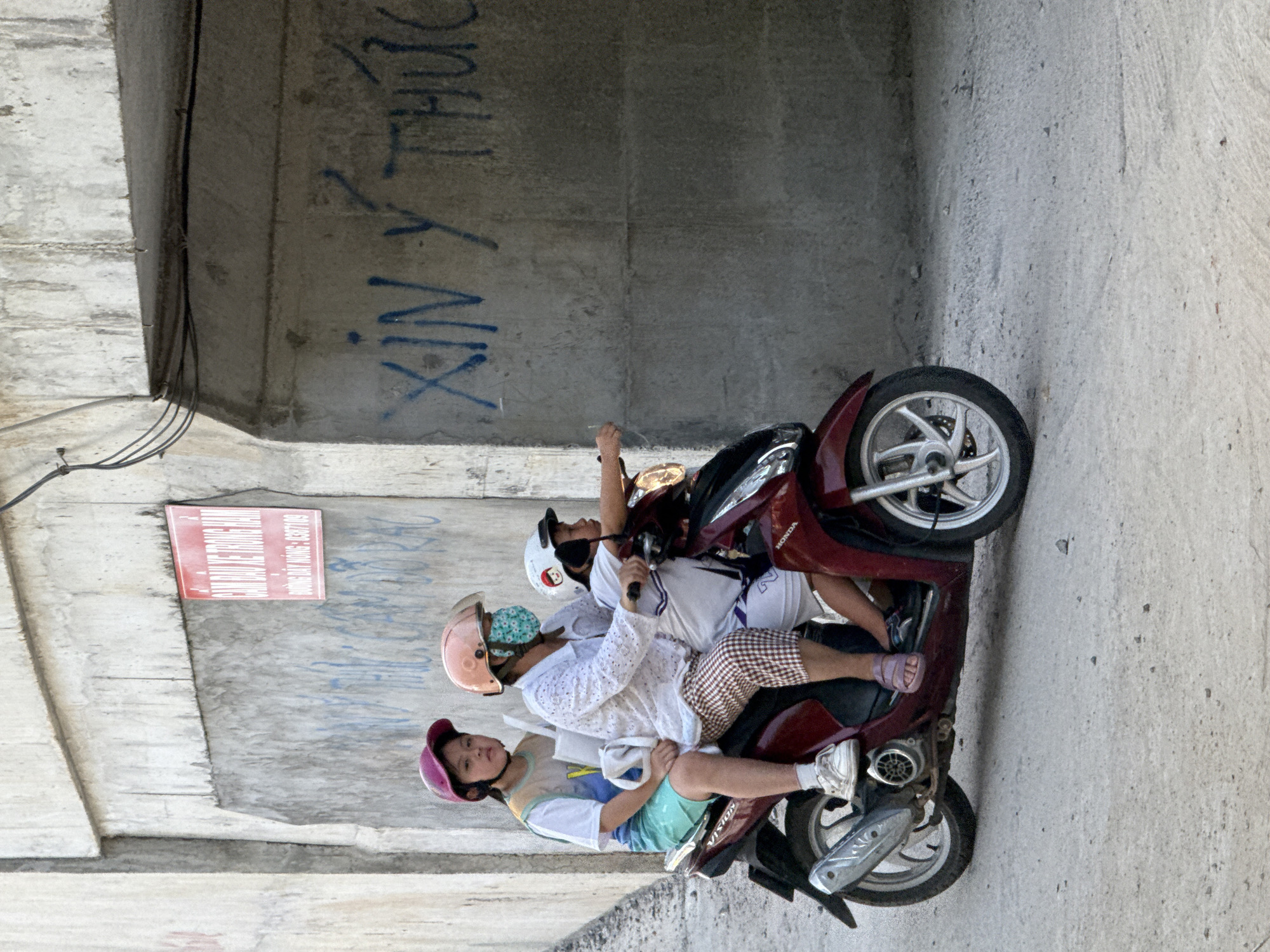 In Bac Nha Trang Ward, Vietnam, three individuals, likely a family, are riding a maroon Honda scooter under a concrete overpass. The adult in the center wears a white shirt and checkered pants, their face obscured by a teal and white patterned mask. To their left, a young girl with a pink helmet wears a white t-shirt with a colorful design and mint green shorts. To the adult's right, a child with a white helmet and a graphic design on their shirt rides in front. The scooter has a silver exhaust pipe and silver rims. The concrete wall behind them is adorned with blue graffiti and a red sign with Vietnamese text. The ground is paved with concrete.