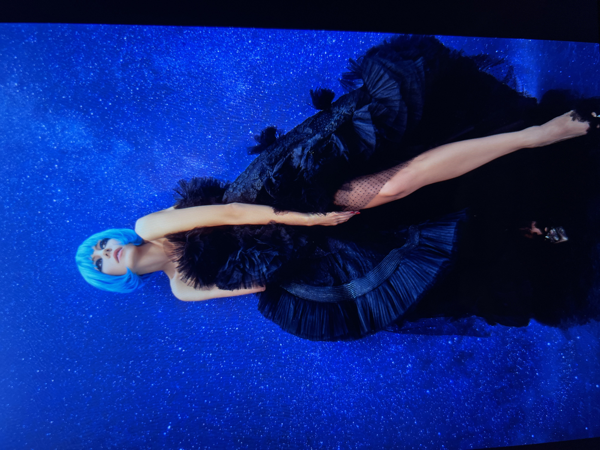 A person with bright blue hair poses against a sparkling blue backdrop. They are wearing a voluminous black gown with intricate ruffles and sheer, polka-dotted fabric on one leg. The person's head is tilted back, and their eyes are looking upwards. The scene is lit dramatically, casting shadows and highlighting the textures of the dress and the sparkle of the backdrop. There is no indication of a specific location in Saint Petersburg, Russia.