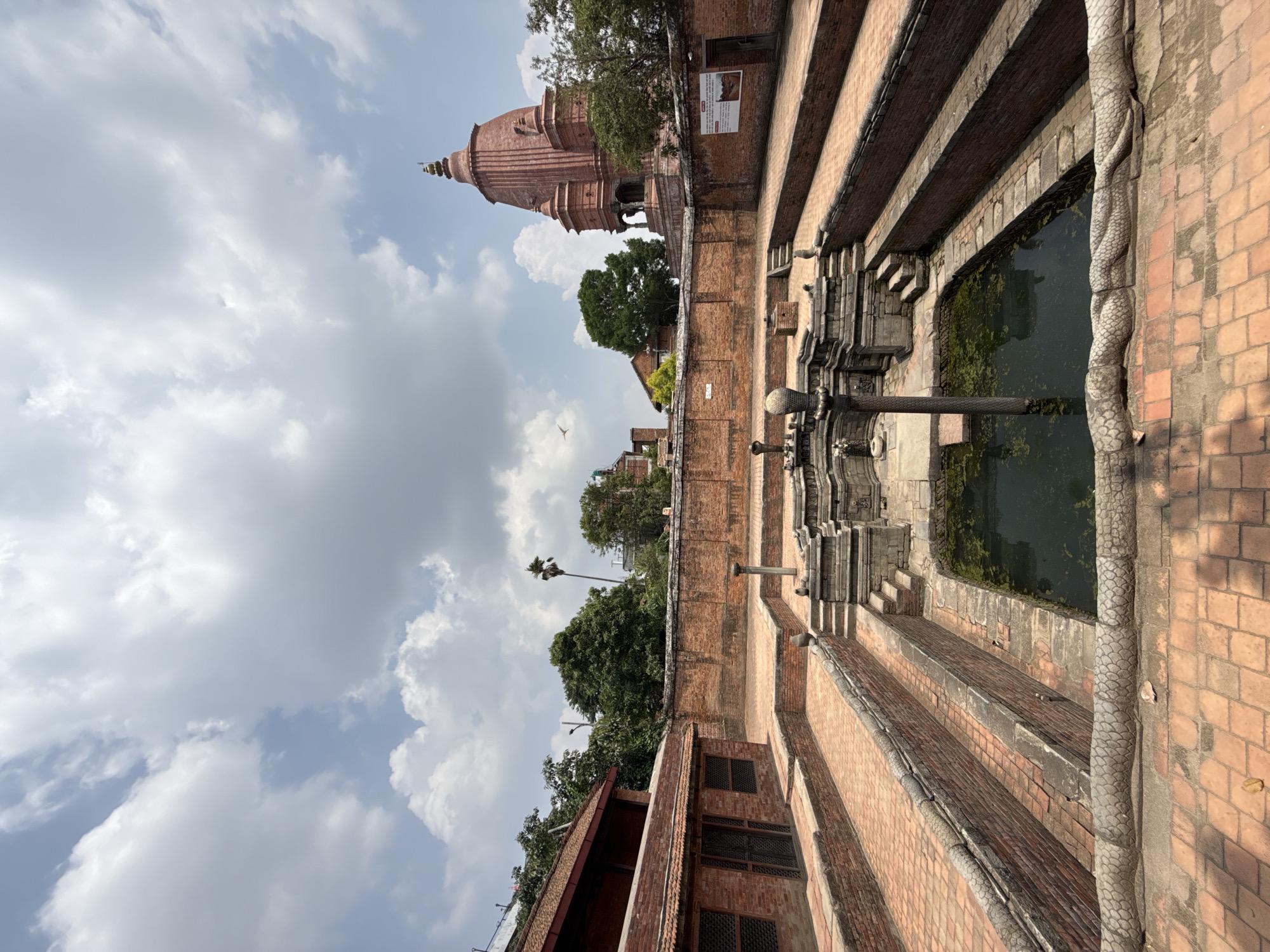 The image depicts the Siddha Pokhari, a historic pond located in Bhaktapur, Nepal. The pond is a rectangular, stepped water tank constructed with brick, filled with green-tinged water. Elaborate brickwork surrounds the pond, including stairs leading down to the water level and decorative brick pillars. A tall, coiled dragon sculpture stands alongside one side of the pond. In the background, a reddish-brown brick temple stands tall amidst green trees against a cloudy sky.