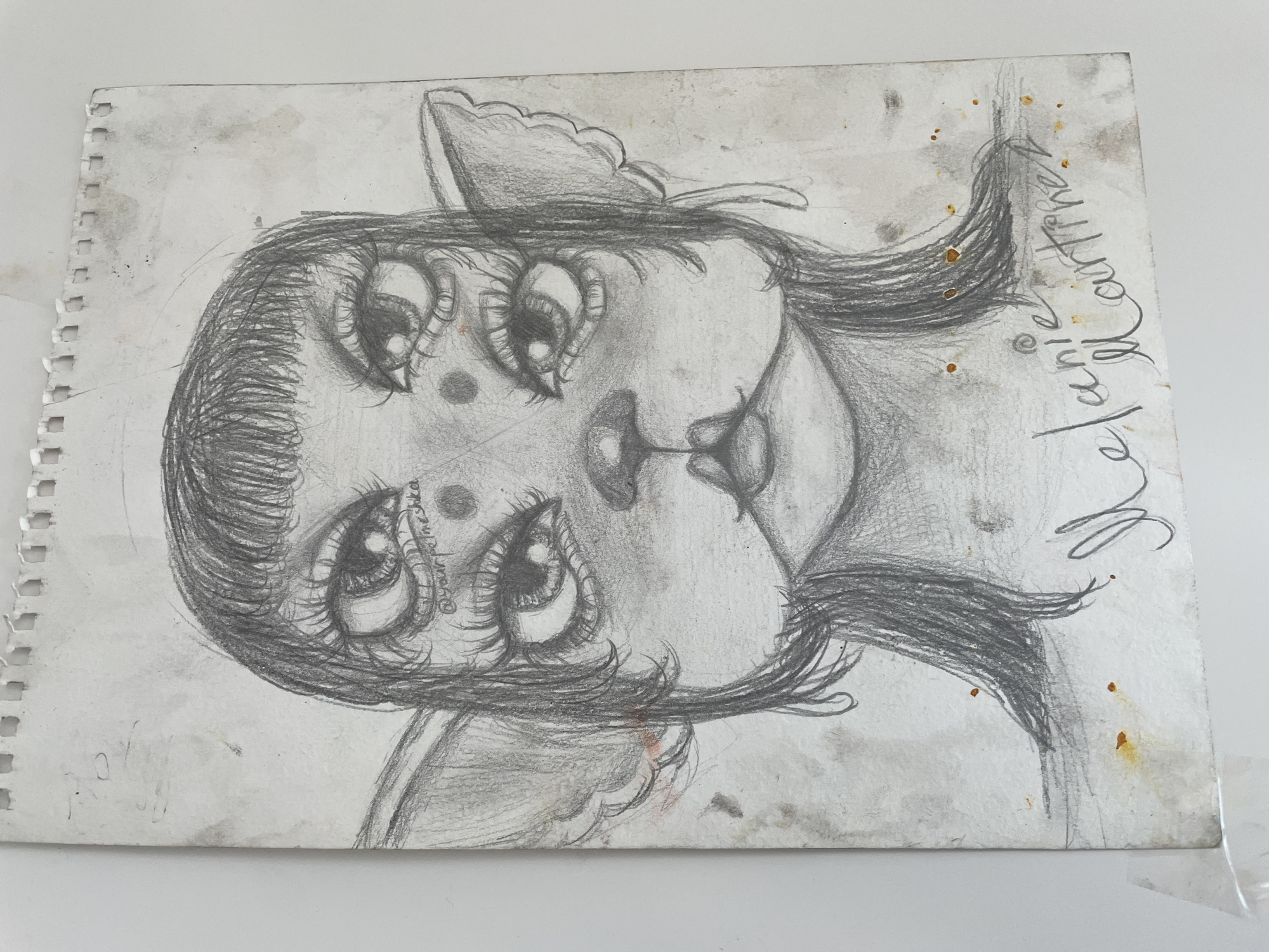 The image is a pencil drawing of a stylized female figure with large eyes and a childlike appearance. The figure has a small nose and mouth, and the hair is depicted with detailed shading. The drawing includes the signature "Melanie lauritzer" and the username "@yourpolmeshka". The paper shows some staining and wear, indicating it is an older or used piece of art. The image itself contains no information relating to the city of Siva, Russia.