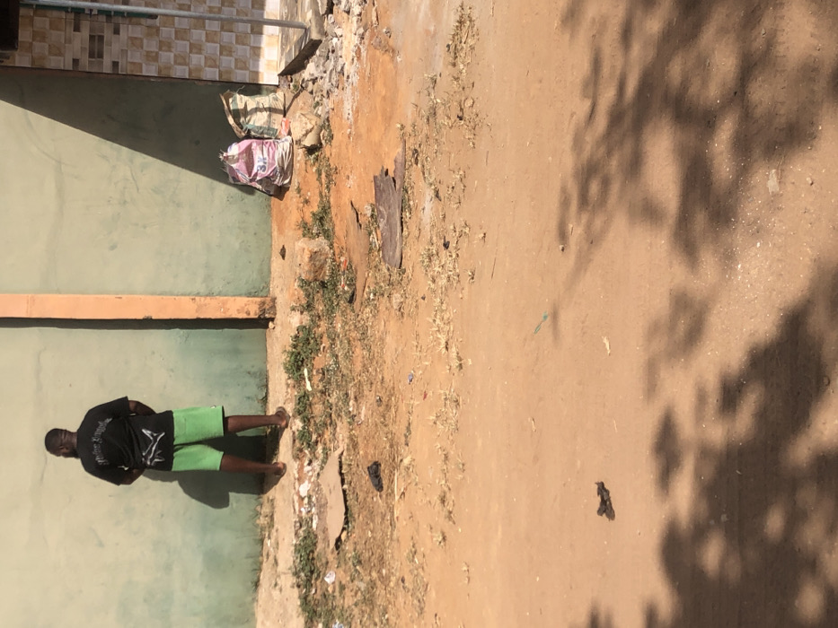 The scene is set outdoors in Egan, Nigeria. A male individual, viewed from the back, stands approximately 0.5 meters from a light green painted wall. He is barefoot, wearing a black t-shirt with a white graphic and bright green shorts. His stance suggests an act of urination or private adjustment. To the right of the individual, the wall transitions to a segment of brown and beige checkered tiles, separated by an orange-brown vertical pillar. Two fabric bags, one pink/white with "3%" visible and another beige/green, rest on the ground near the tiled section. The ground consists of reddish-brown dirt, sparsely covered with dry green vegetation and scattered debris including cardboard pieces, dark plastic, and concrete fragments. Strong daylight is indicated by distinct shadows cast by the individual on the wall and by unseen foliage in the foreground.