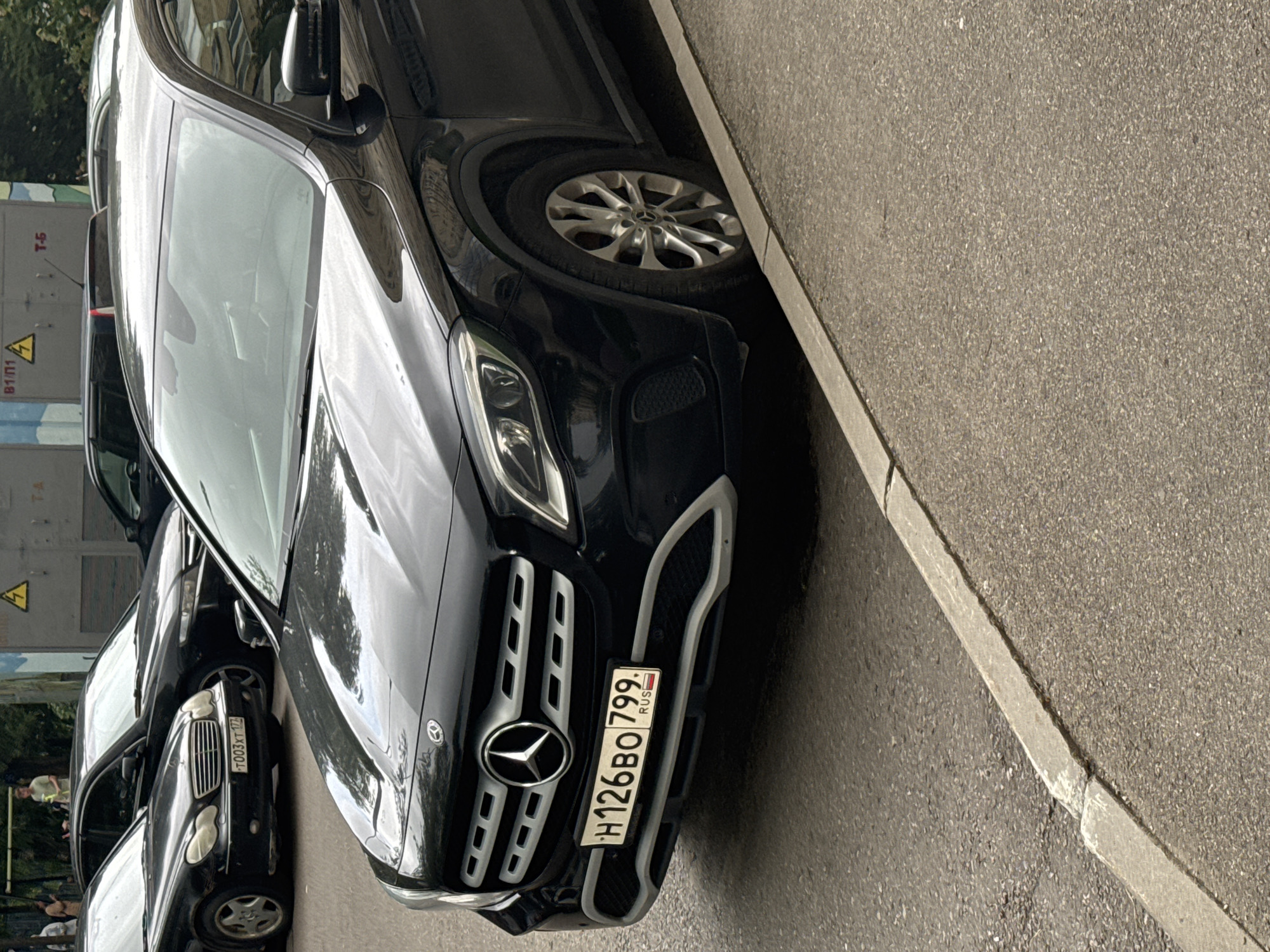 In Zelenograd, Russia, a black Mercedes-Benz CLA-Class is parked on an asphalt road next to a concrete curb. The vehicle's license plate reads "H126BO 799". To the left of the Mercedes, a black Mercedes-Benz E-Class is partially visible. In the background, people are standing near a building with electrical equipment and warning signs. The scene appears to be a typical urban street.
