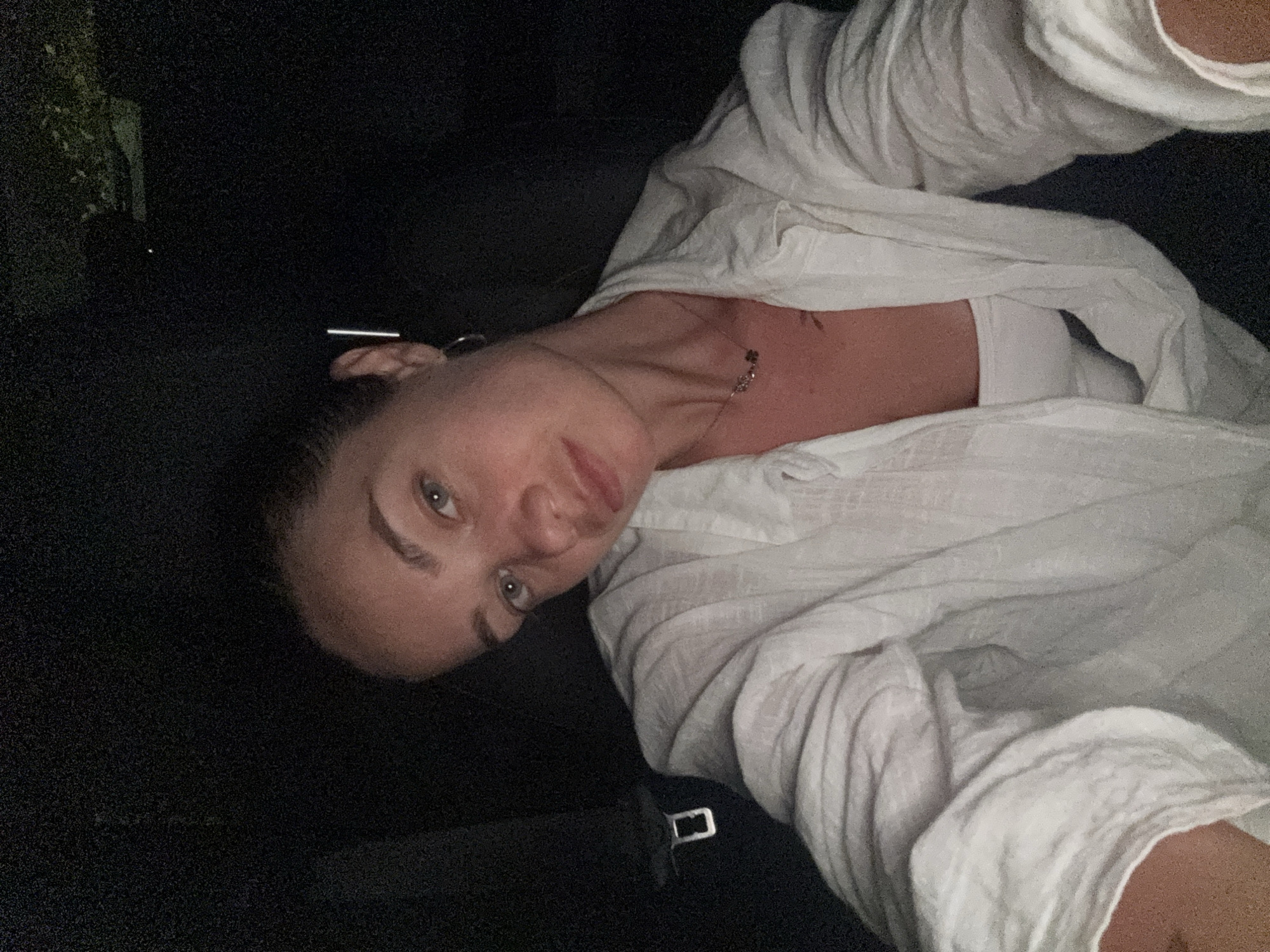 A young Caucasian woman with fair skin and light brown hair pulled back is shown in a low-light, close-up selfie. She is wearing a white, open, linen-like button-down shirt over a white tank top, and a delicate necklace with small charms. A faint tattoo is visible on her upper chest. She has a silver bar hairpin in her hair and a hoop earring in her visible ear. The background is dark, suggesting an indoor setting at night, possibly a car interior given the presence of a seatbelt buckle visible on the left side of the frame. The image is taken at a slightly upward angle, focusing on her face and upper body. The woman's gaze is directed towards the camera with a neutral expression. The location is specified as Radovići, Montenegro.