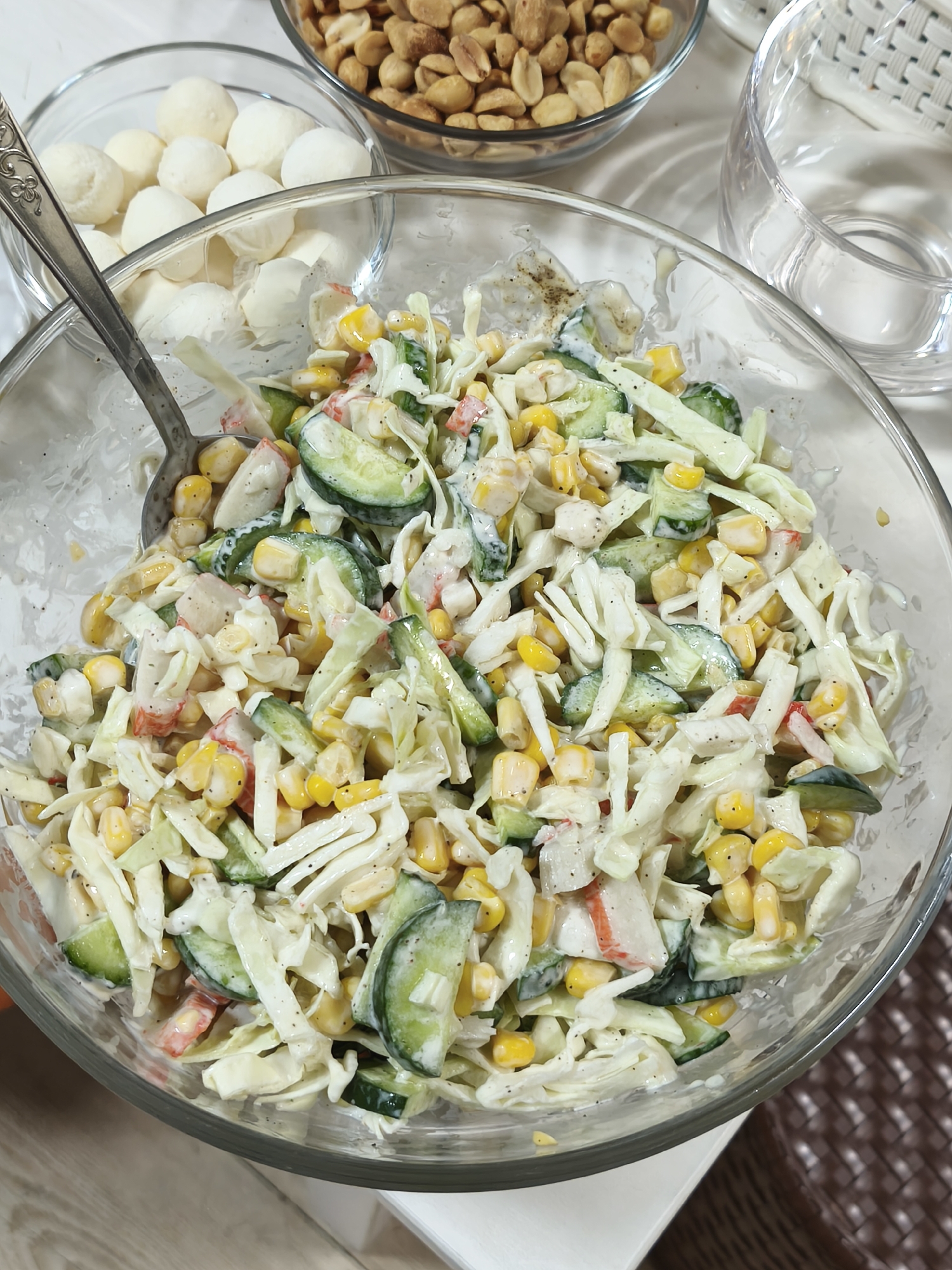 A close-up, overhead shot shows a clear glass bowl filled with a salad. The salad contains shredded cabbage, corn kernels, sliced cucumbers, and pieces of imitation crab meat, all coated in a creamy dressing. A silver serving spoon rests in the bowl, partially submerged in the salad. In the background, another clear bowl holds roasted peanuts. To the right of the salad bowl, a clear glass is visible. The setting appears to be a table, with a white surface in the foreground and a textured brown placemat visible in the lower right corner. No specific location in Asan-si, South Korea can be determined from the image.