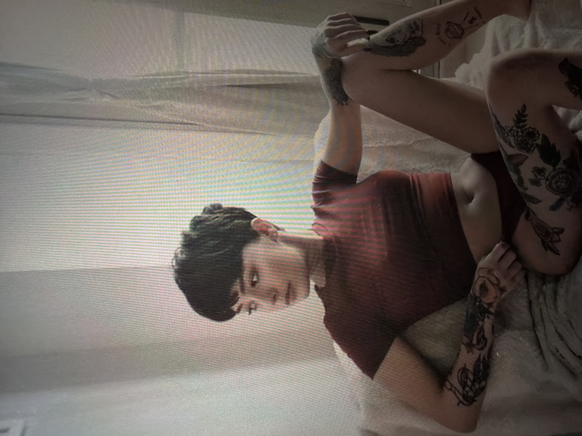 A young woman with short, dark hair and visible tattoos on her arms and legs is seated on a couch. She is wearing a short-sleeved red top and dark bottoms. The environment appears to be indoors, with sheer curtains in the background and diffused light. There are no discernible Shumen, Bulgaria landmarks present in the image.