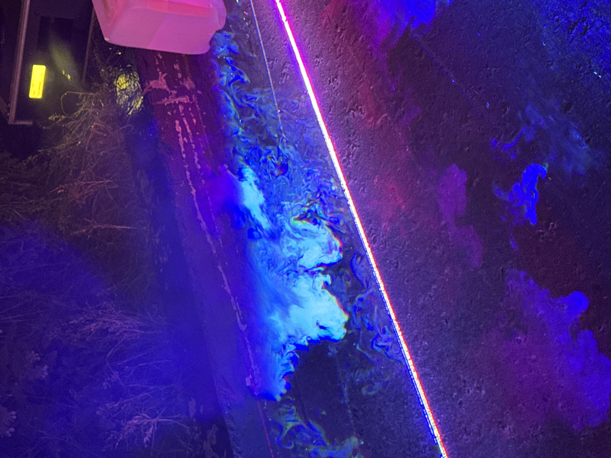 The image displays an outdoor, nighttime scene illuminated by diverse artificial lighting. The ground surface, appearing dark and textured, possibly asphalt or concrete, occupies the lower and mid-sections of the frame. Across the mid-ground, a prominent, straight, luminous strip, displaying red and blue light components, extends diagonally from the lower left towards the upper right.
To the upper left of this light strip, a substantial volume of bright, light blue-cyan material, possibly a fluorescent liquid or dense smoke, swirls and spreads across the dark ground. This material exhibits intricate, organic patterns and integrates with subtle green and yellow hues. Directly above the right end of the light strip, a partially visible, translucent, rectangular object, glowing pinkish-red, stands vertically at the far right edge of the frame. Further back, the ground shows rough textures and lighter streaks under pink-purple illumination.
The background on the left is dominated by dense, dark vegetation, including bushes and possibly small trees, bathed in deep blue and purple light. On the right side of the background, an unidentified dark structure is partially visible behind the vegetation, featuring a distinct, bright yellow rectangular light or reflector. The overall lighting scheme is characterized by intense blue, purple, pink, and yellow artificial lights, suggesting the use of UV or specialized colored illumination. No individuals or animals are present in the image. The image contains no discernible information or landmarks to indicate the specific location within the city of Moscow, Russia.