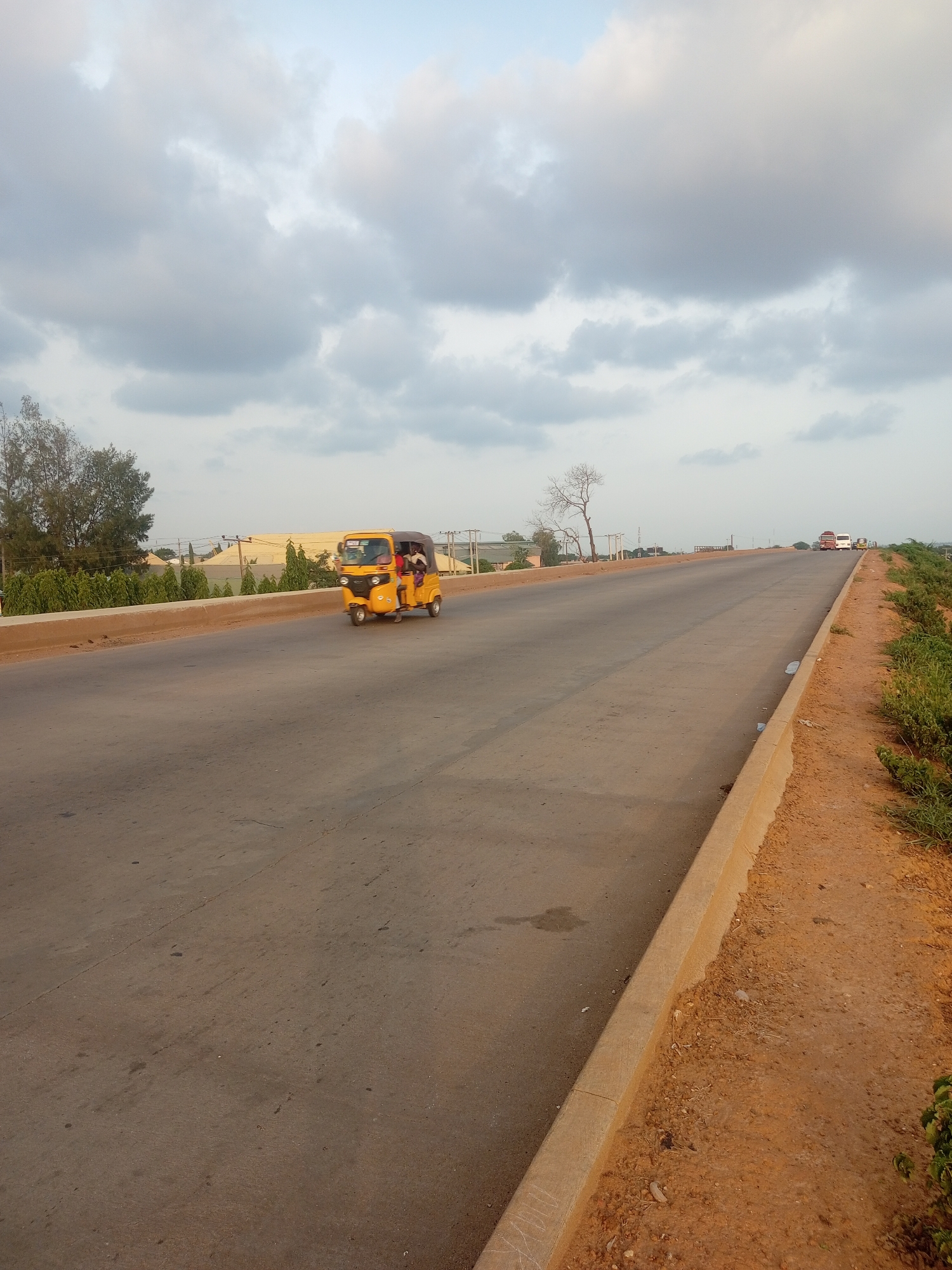 The image depicts a bright yellow, three-wheeled auto-rickshaw driving on a paved road under a cloudy sky. The auto-rickshaw contains at least two passengers. The road is wide and appears to be an elevated highway, with a concrete barrier separating it from a dusty embankment on the right side, which is covered in sparse green vegetation. In the background, there are trees, some buildings with light-colored roofs, and electrical poles. Further in the distance, a few other vehicles, including what appear to be buses, are visible. The overall scene suggests an outdoor setting on a road, possibly in a semi-urban or rural area.