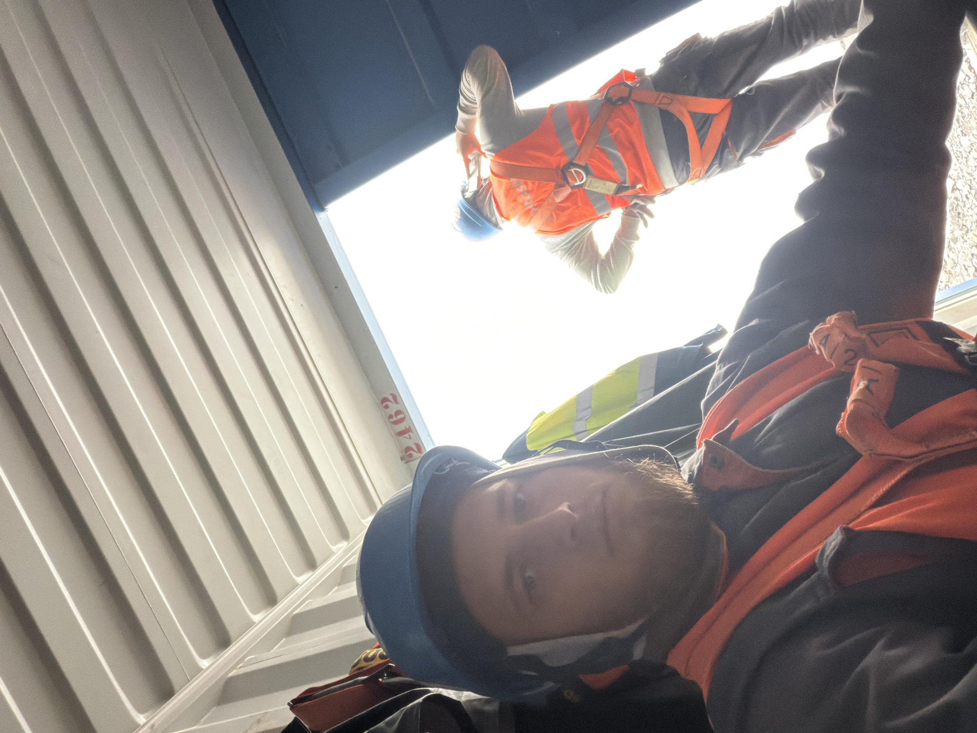 The image shows two construction workers inside a shipping container, with the doors open to a bright outdoor environment. The worker in the foreground is wearing a blue hard hat, an orange high-visibility vest over a dark grey jacket, and a harness. They are looking directly at the camera with a serious expression and have a beard. The second worker, in the background, is also wearing a blue hard hat and an orange high-visibility vest with a harness, and appears to be on their phone. The interior of the container has a corrugated metal ceiling. A red number "2462" is visible on the ceiling towards the rear of the container. The overall lighting suggests it is daytime. There is no information in the image to suggest the location is in Janowice, Poland.