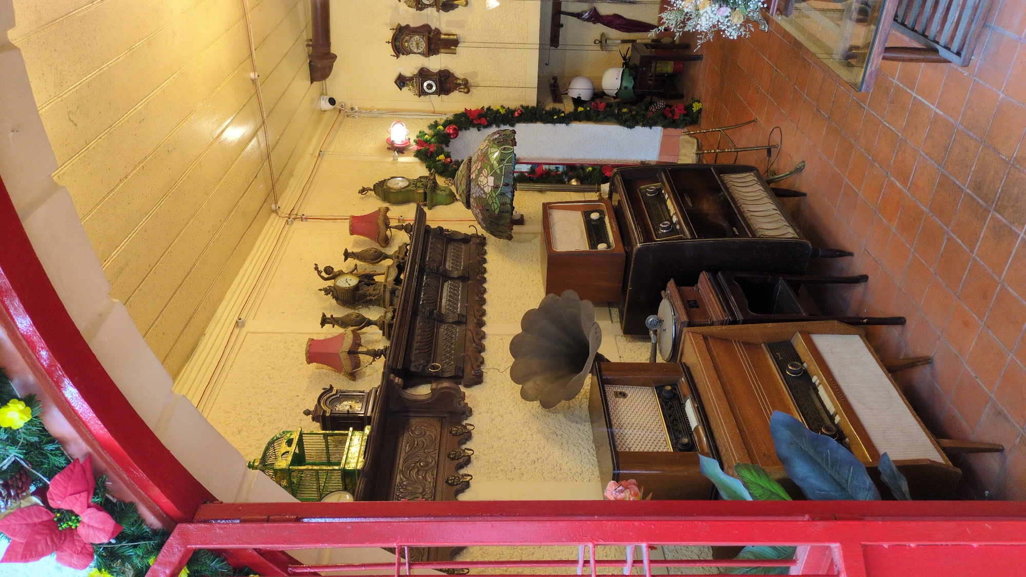 The image depicts the interior of a shop in Da Lat, Vietnam, filled with vintage items. The scene is dominated by antique radios, gramophones, clocks, and lamps, suggesting a collection of historical artifacts. A red-painted archway frames the left side of the image, adorned with festive greenery and red and yellow flowers, possibly indicating a holiday season. The walls are textured and painted a light yellow. The floor is tiled with terracotta squares. Several wooden furniture pieces, including cabinets and stands, hold the displayed objects. In the background, a decorative garland with red bows and pinecones hangs near an arched doorway, leading to another area of the shop. Various clocks are visible on the wall, and a stained-glass lamp with floral motifs stands prominently. The overall impression is that of a dimly lit, cluttered space dedicated to showcasing nostalgic memorabilia.