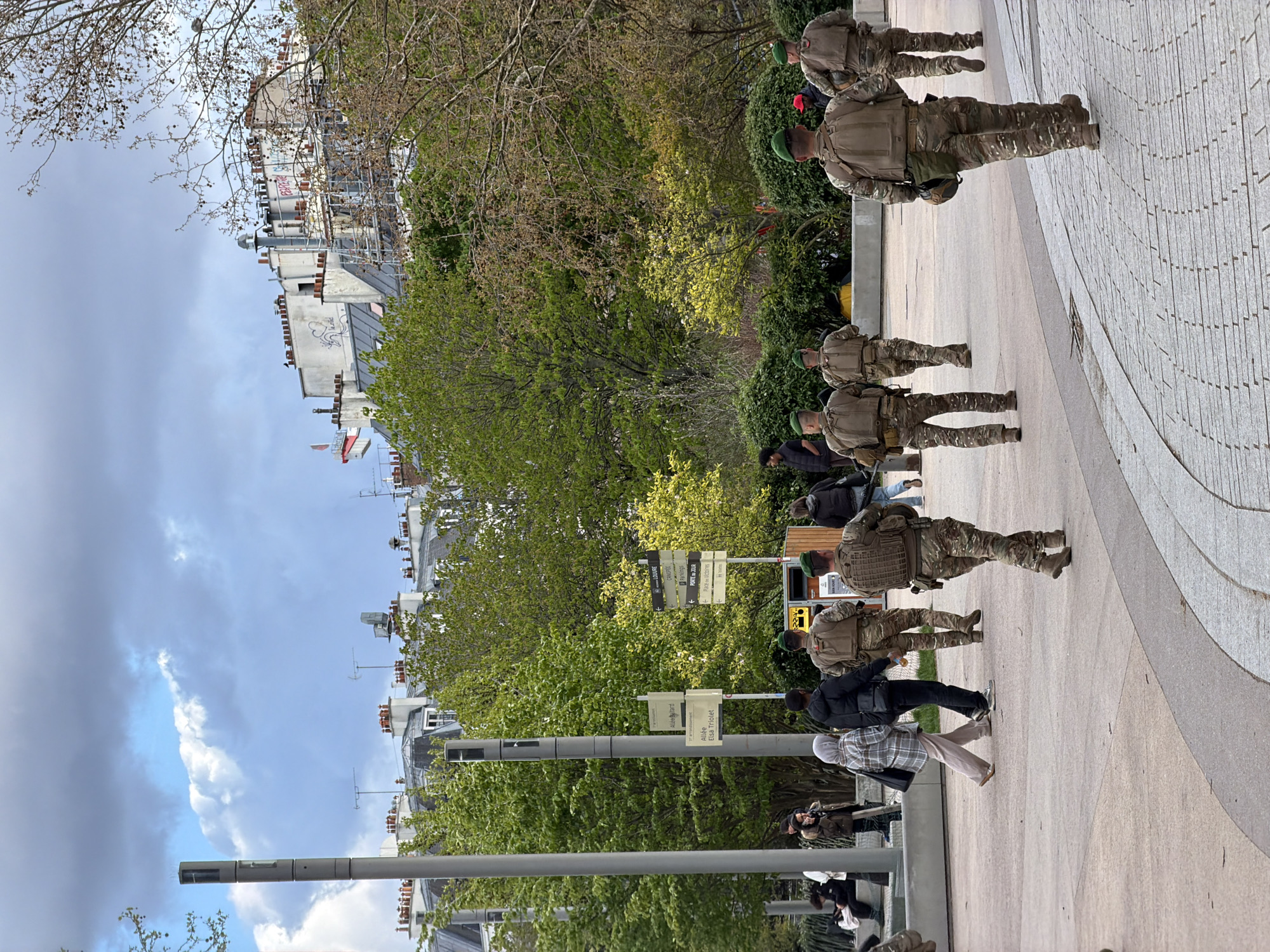 This image captures an urban pedestrian area within the Forum des Halles complex in Paris, France, specifically indicated by signs for "Allée Elsa Triolet," "Allée Ballard," and "3e arrondissement," situated near the border of the 1st and 3rd arrondissements. The scene is observed under a partly cloudy sky.
In the mid-ground and foreground, a group of at least six individuals, presumed to be military personnel, walk away from the viewer. They wear camouflage fatigues, tactical plate carrier vests, and green berets. The uniform pattern appears to be multi-cam. One soldier on the far right has an obscured object, possibly a weapon, slung across his back. They move along a paved pathway composed of light-colored concrete and darker grey stone pavers.
Numerous civilians are also present, occupying various parts of the scene. One woman in a light-colored plaid jacket, light trousers, and a hijab walks alongside a man in a dark jacket and dark trousers. Other civilians are visible further back, some seated on ledges or walking near trees.
The environment features mature green trees and bushes, some exhibiting nascent spring foliage. Dominating the background are traditional Parisian buildings with light-colored facades, dark mansard roofs, and numerous chimneys. Graffiti, including the word "PARIS" in red, is visible on upper sections of some buildings. Scaffolding is present on one building's facade. Modern grey cylindrical urban poles, possibly streetlights or surveillance fixtures, are prominent. Several informational signs provide directions to local landmarks such as "LOUVRE" and "CHATELET." A yellow ticketing or information kiosk is also visible.