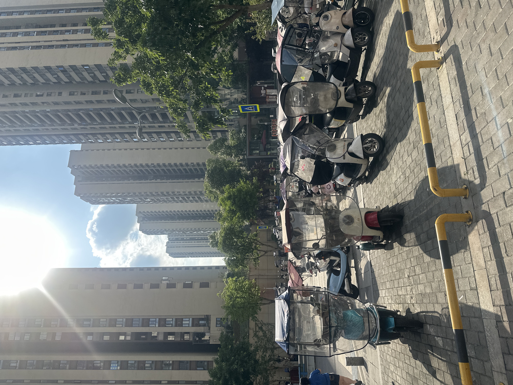 In the city of Chongqing High-tech Zone, China, a row of electric scooters with clear plastic covers is parked on a brick-paved surface. The scooters are of various colors, including teal, red and white, and white. Some have transparent roofs and windshields. Behind the scooters, several tall, modern apartment buildings rise into the sky, with the bright sun casting long shadows across the scene. A few trees are visible, adding a touch of green to the urban landscape. In the foreground, yellow and black striped bollards delineate a walkway. A person wearing a blue top and dark shorts is walking away from the camera on the left side of the image. The sky is bright blue with some fluffy white clouds.