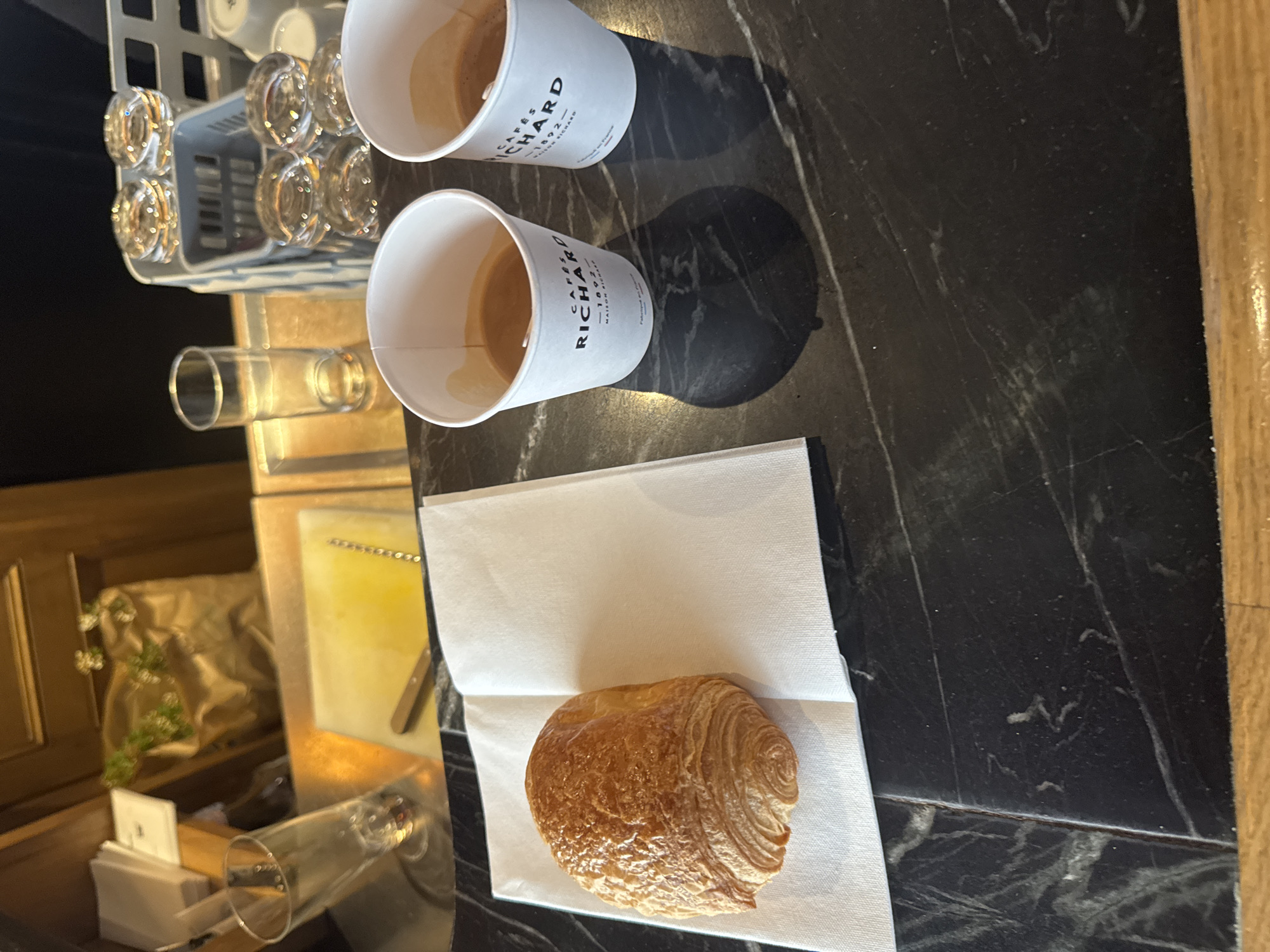 The image captures a scene likely in a Parisian café. On a dark, marble-patterned tabletop, there are two paper cups labeled "CAFÉS RICHARD," both partially filled with coffee. Beside them lies a folded white paper napkin, upon which a golden-brown croissant rests. In the background, a dish rack filled with glassware is visible, alongside a serving tray and some decorative items. The wooden paneling suggests an indoor setting, possibly inside a cafe.