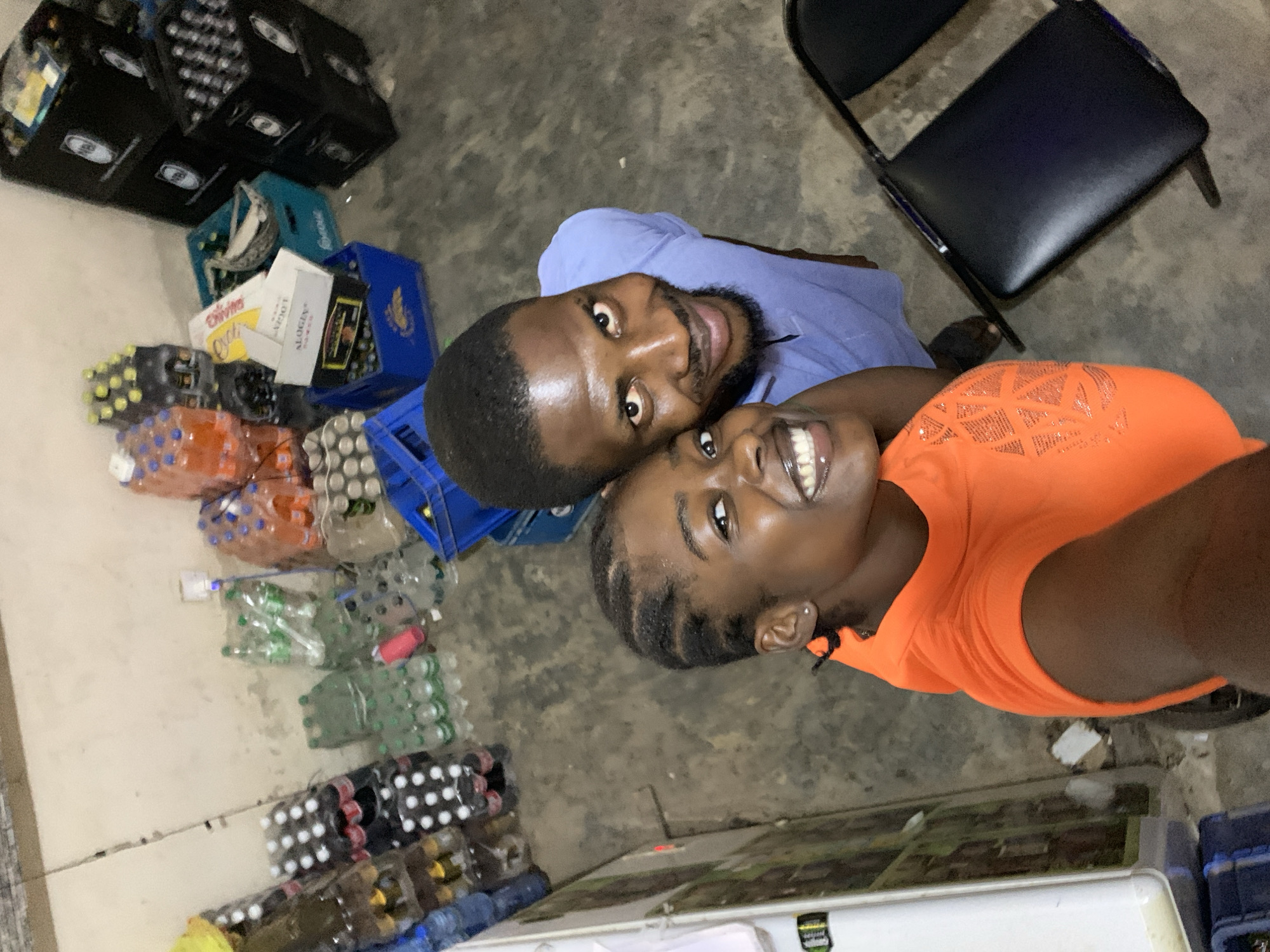 The image shows two people, a man and a woman, smiling at the camera. They are standing in what appears to be a storage or warehouse type of room in New Stadium, Nigeria. The man is wearing a light blue shirt and the woman is wearing an orange shirt. There are numerous stacks of bottle crates, some full of drinks, lining the walls. A black chair is visible in the upper right corner. The room has a concrete floor.