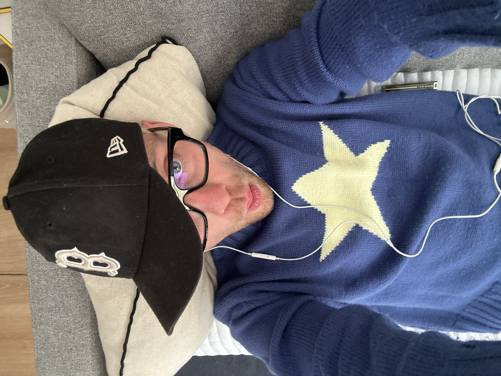 A fair-skinned male, presumably an adult, is lying on a gray couch or sofa. He's wearing a black baseball cap with a white "B" logo on the side, black-framed glasses, and a blue sweater adorned with a large yellow star. White earbuds are plugged into his ears. A beige pillow with dark trim is beneath his head. There's no indication of this image being from London or the United Kingdom; the setting appears to be indoors, but the specific location cannot be determined.
