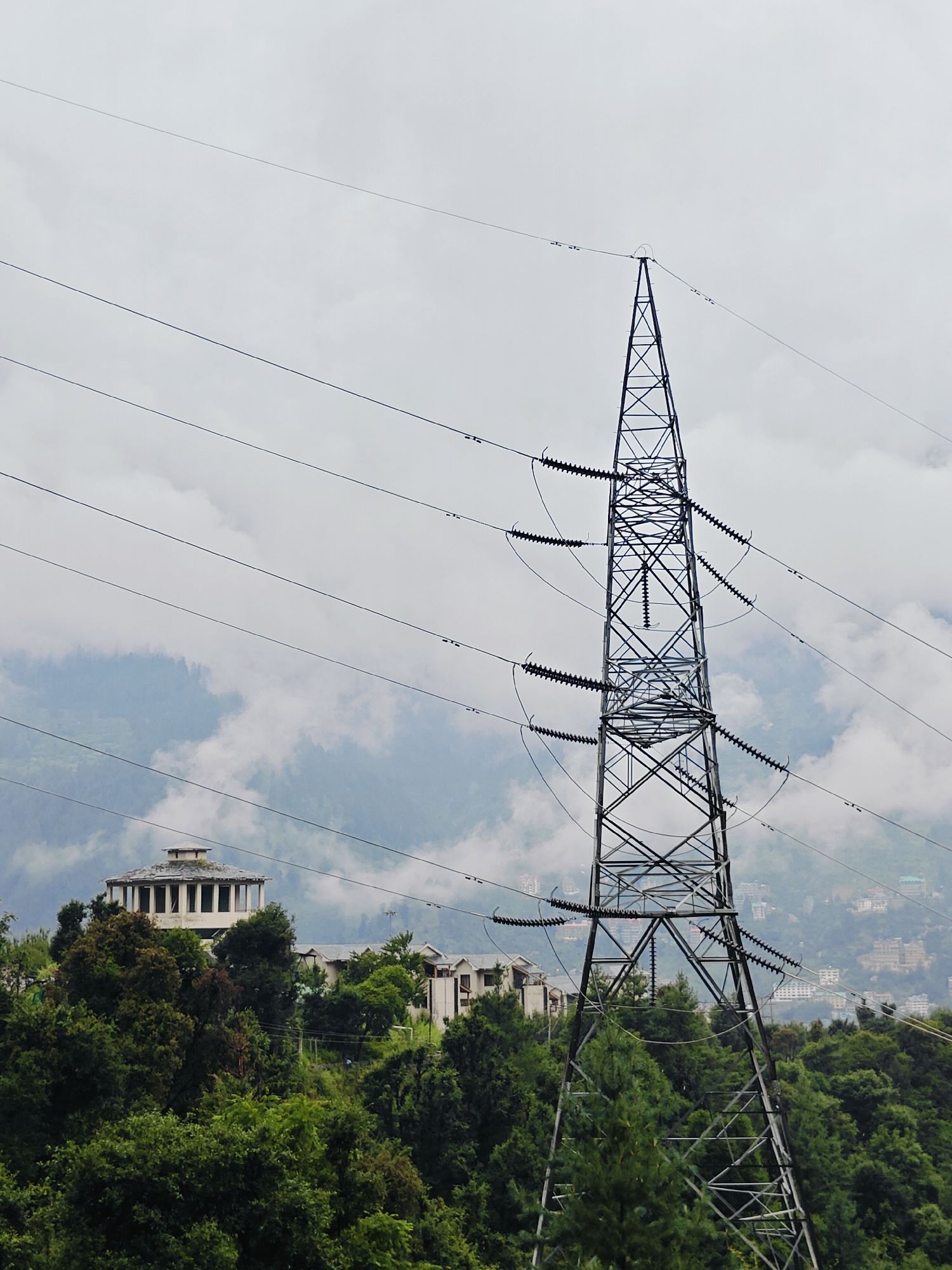In Prini, India, a large, dark metal electricity pylon stands tall in the foreground, its lattice structure reaching towards a cloudy sky. Several thick, dark electrical wires extend horizontally across the frame, suspended from the pylon and disappearing into the distance. In the mid-ground, nestled amongst lush green trees and rolling hills, a distinctive circular building with a flat roof and numerous evenly spaced windows is visible. Adjacent to it, other buildings with white facades and pitched roofs are partially obscured by the dense foliage. The background reveals a hazy, mountainous landscape, with a town or village spread across the slopes, its buildings appearing as small white and colored dots against the muted greens and grays of the terrain. The overall atmosphere is overcast, with soft light and a sense of depth created by the layers of landscape.